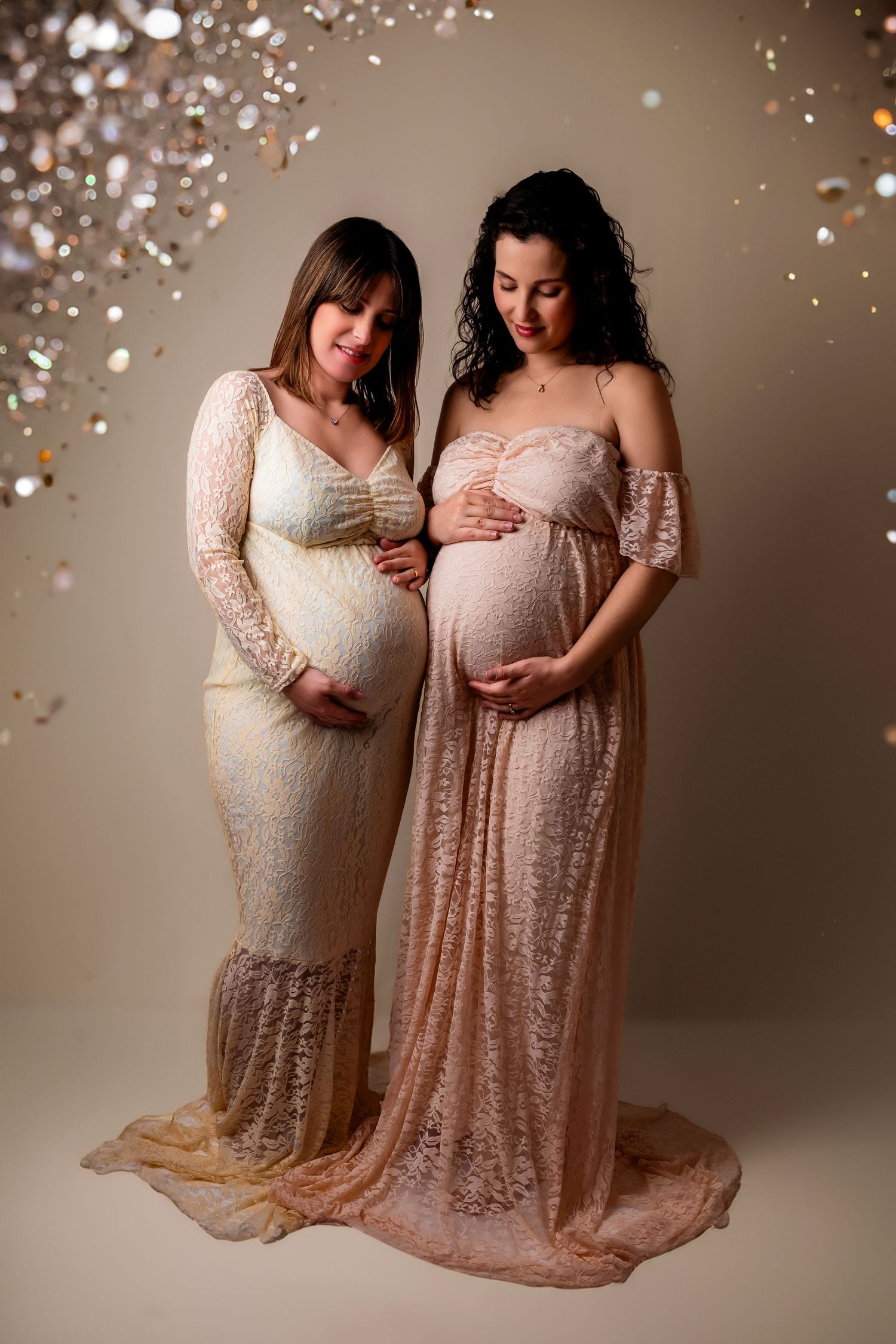 Two pregnant women in lace maternity gowns pose together as glittering confetti falls around them in a studio setting.