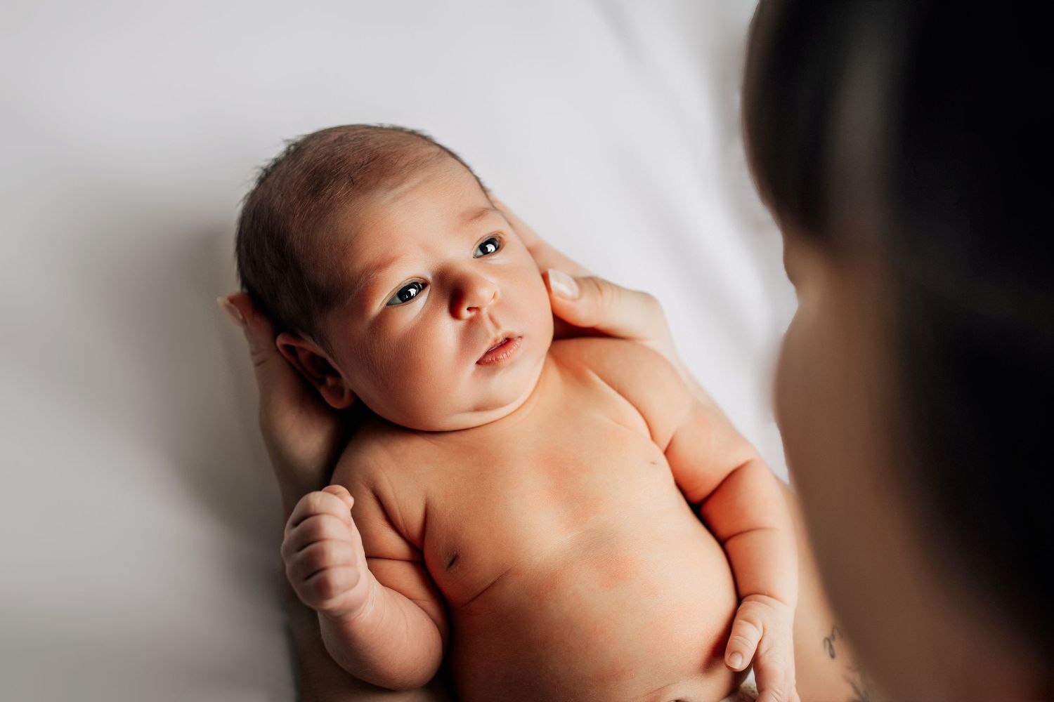 A newborn baby lies on white bedding while looking upward with soft natural lighting.