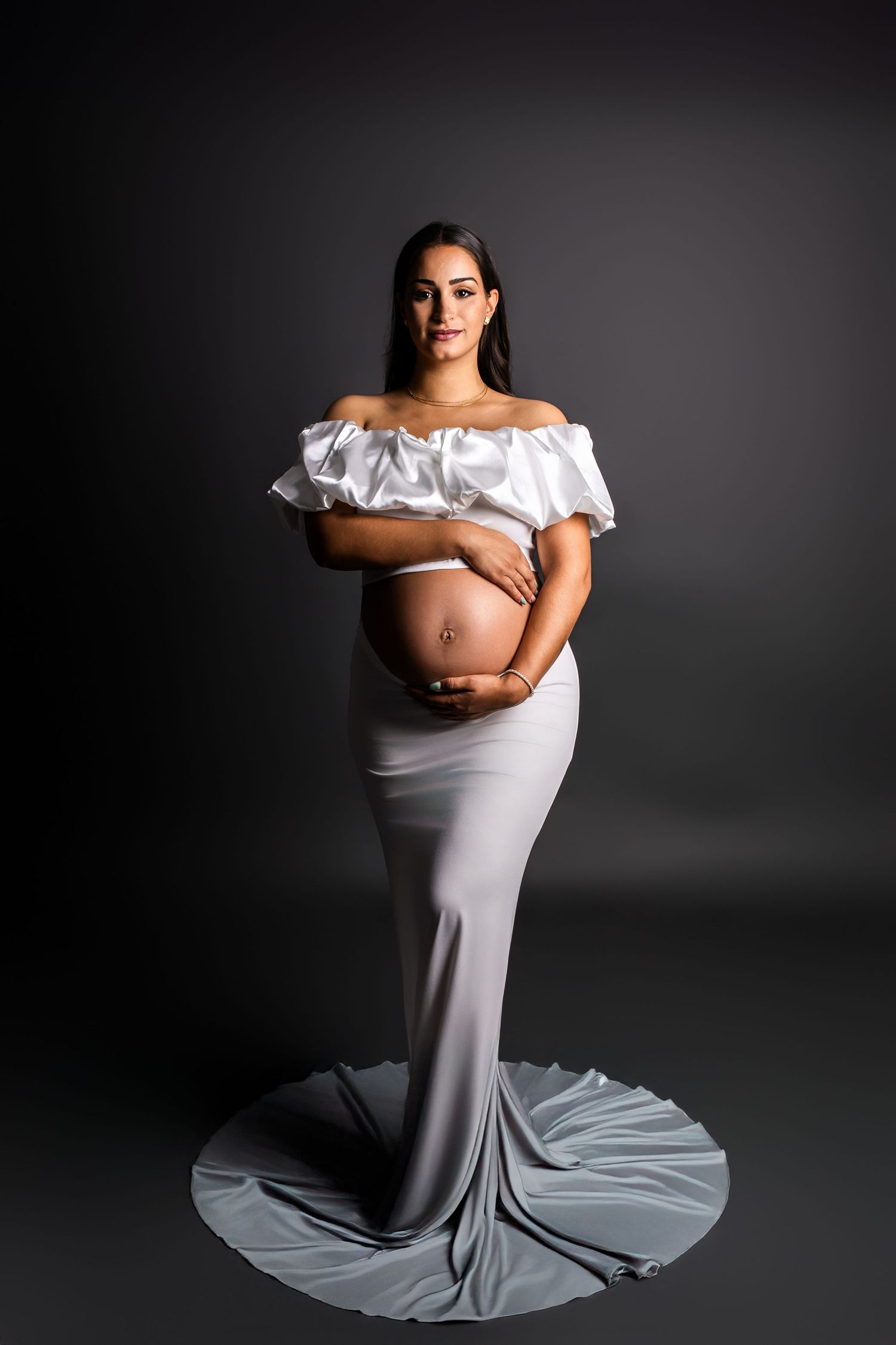 A dramatic maternity photoshoot series in black and white featuring a woman in a flowing white gown against dark backdrop.