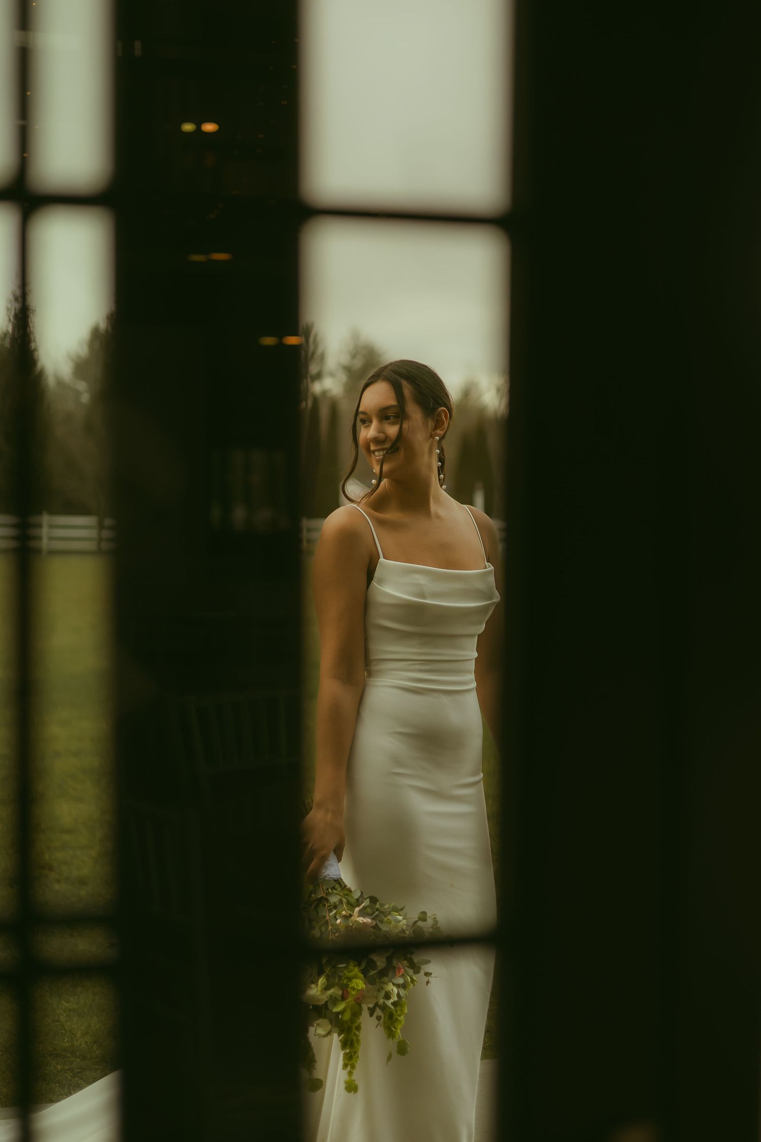 A bride in a sleek white dress seen through window panes holding a bouquet.