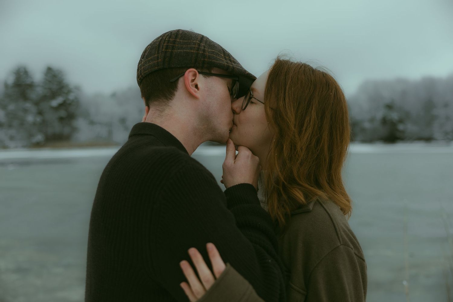 A couple shares a tender kiss outdoors on a misty day with evergreen trees visible in the background.