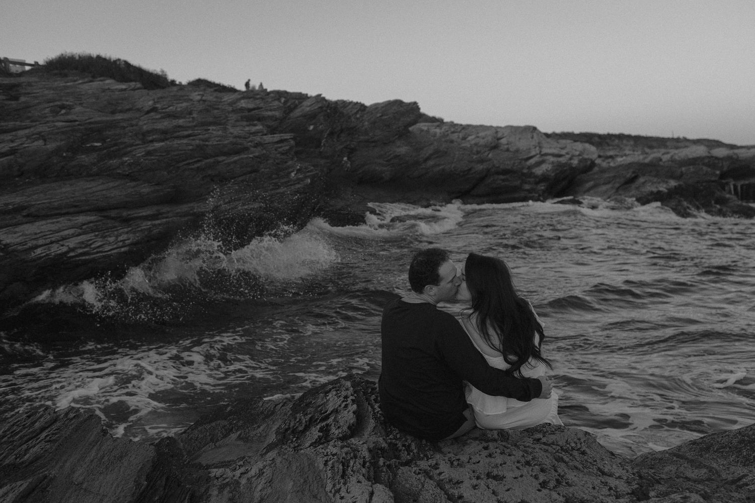 People exploring rocky coastal cliffs at sunset with ocean waves crashing below.