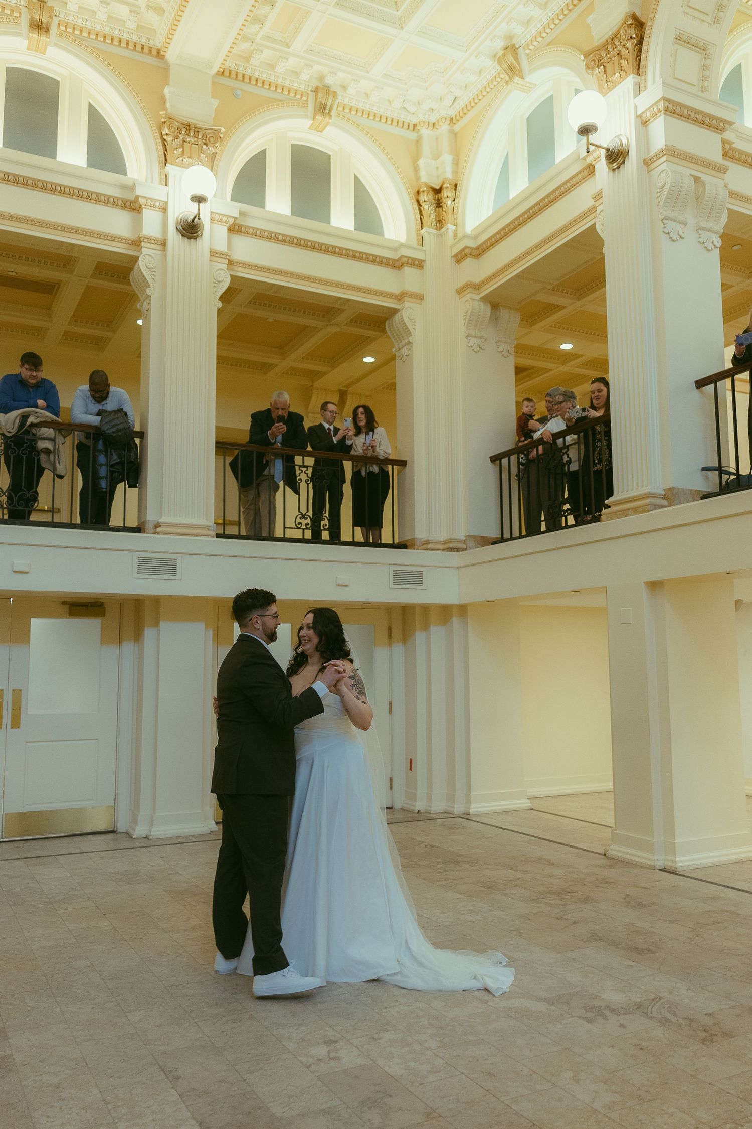 Elegant wedding couple dances in a grand ballroom with ornate white columns and balcony overlooking the scene.