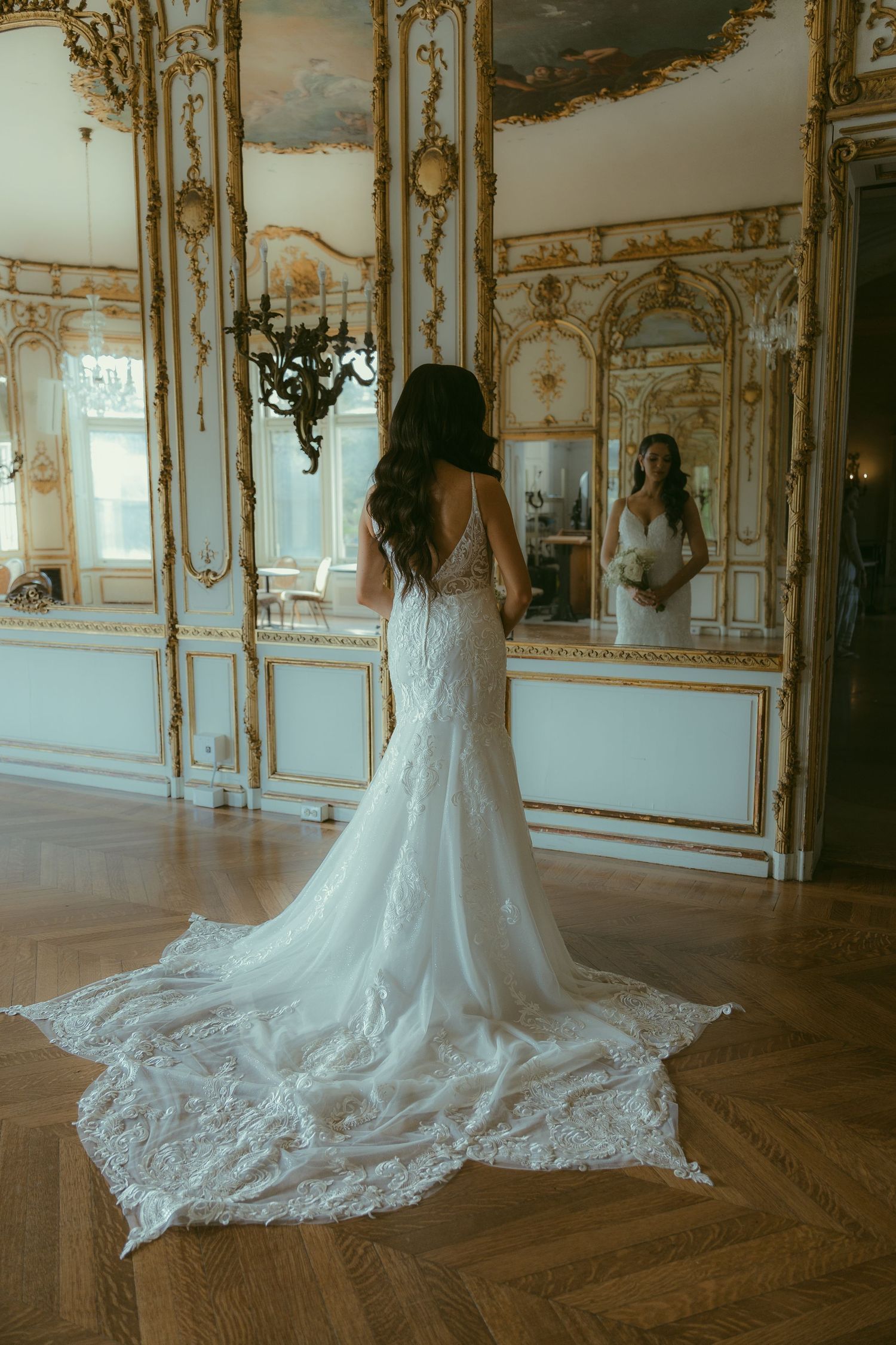 A bride in a lace wedding dress with a long train stands in an ornate mirrored ballroom.