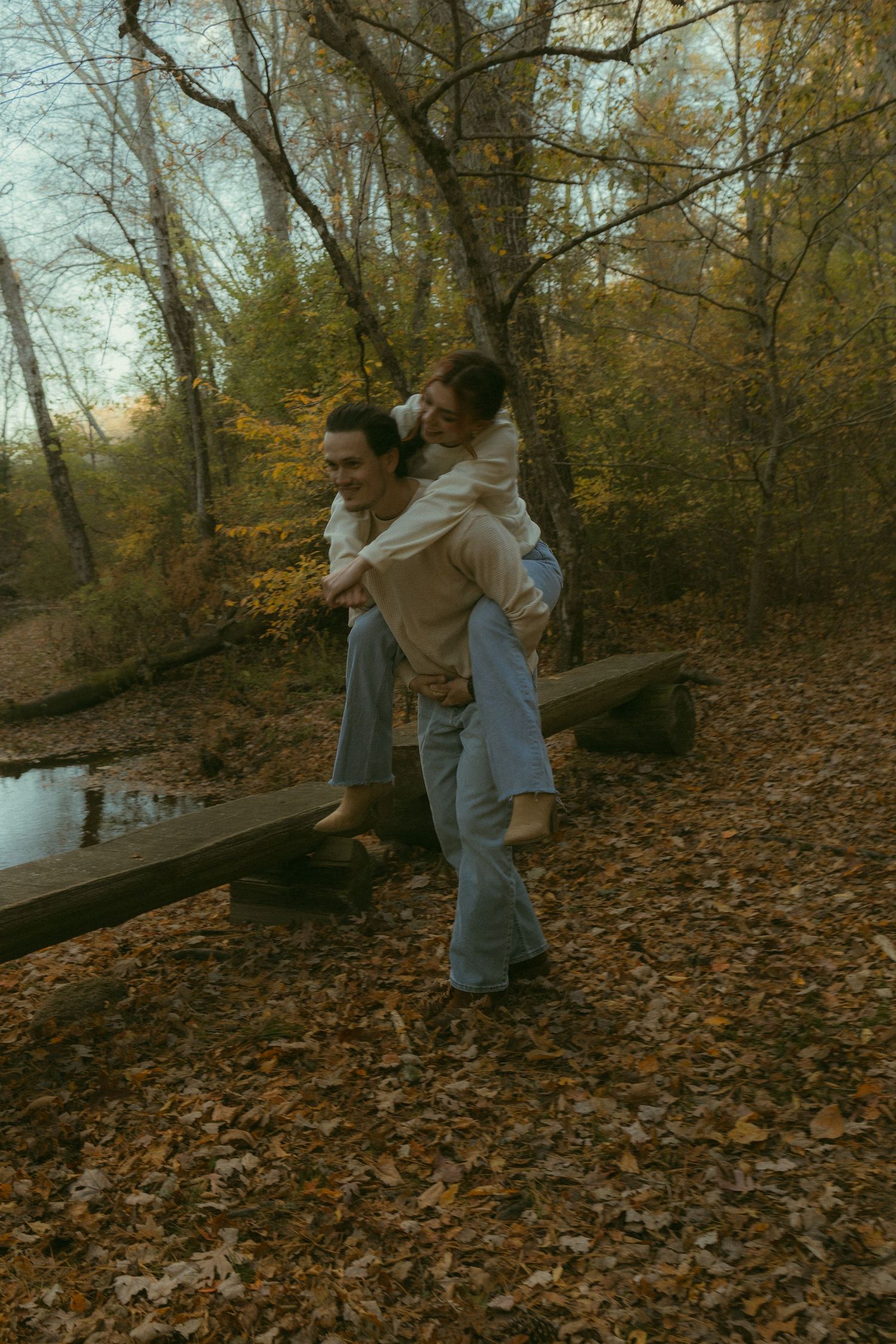 Couple playfully dancing and embracing among fallen autumn leaves in a wooded park setting.