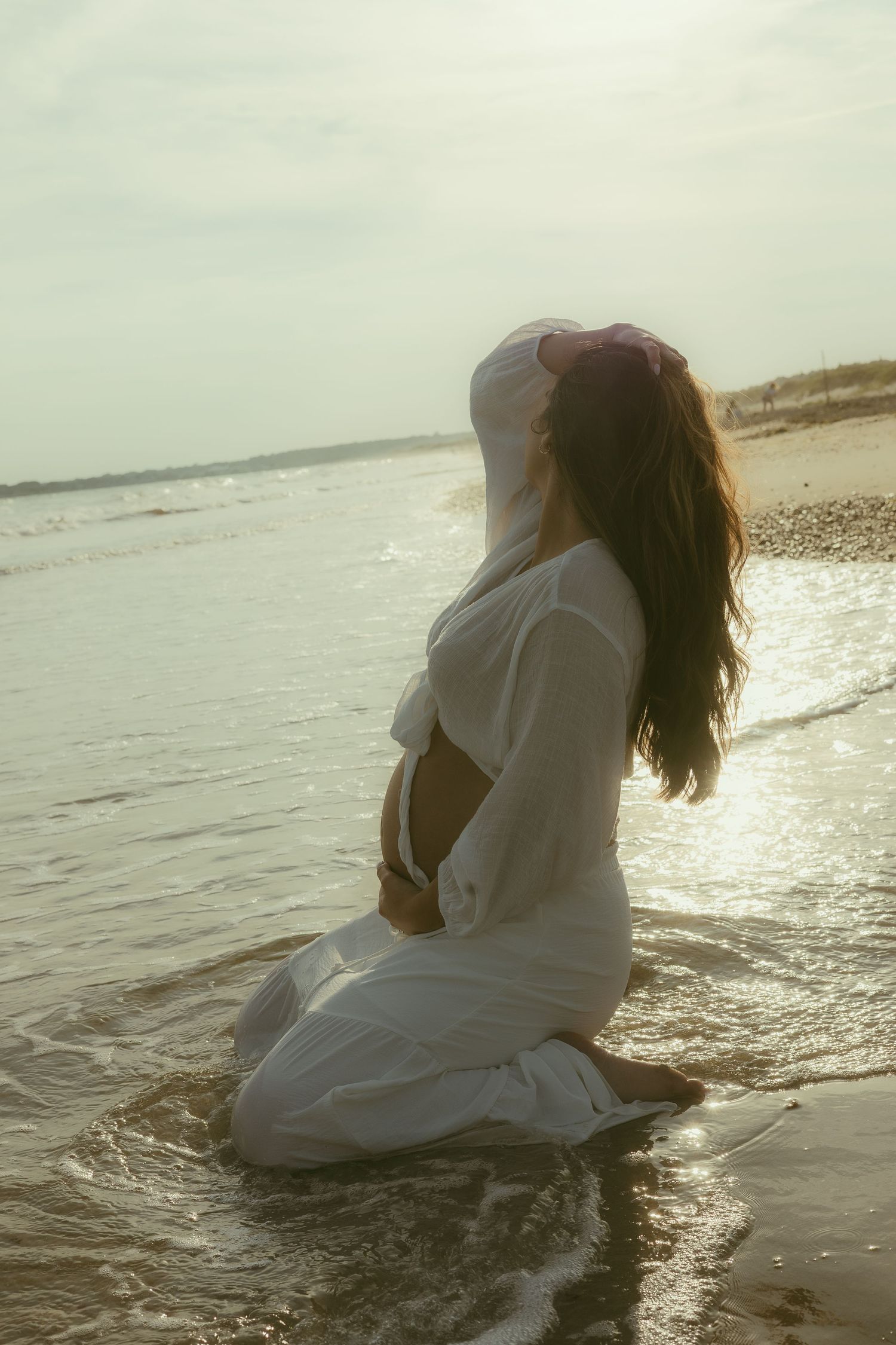 Two figures in white clothing embrace on a beach shoreline at sunset creating a romantic silhouette against the ocean waves.