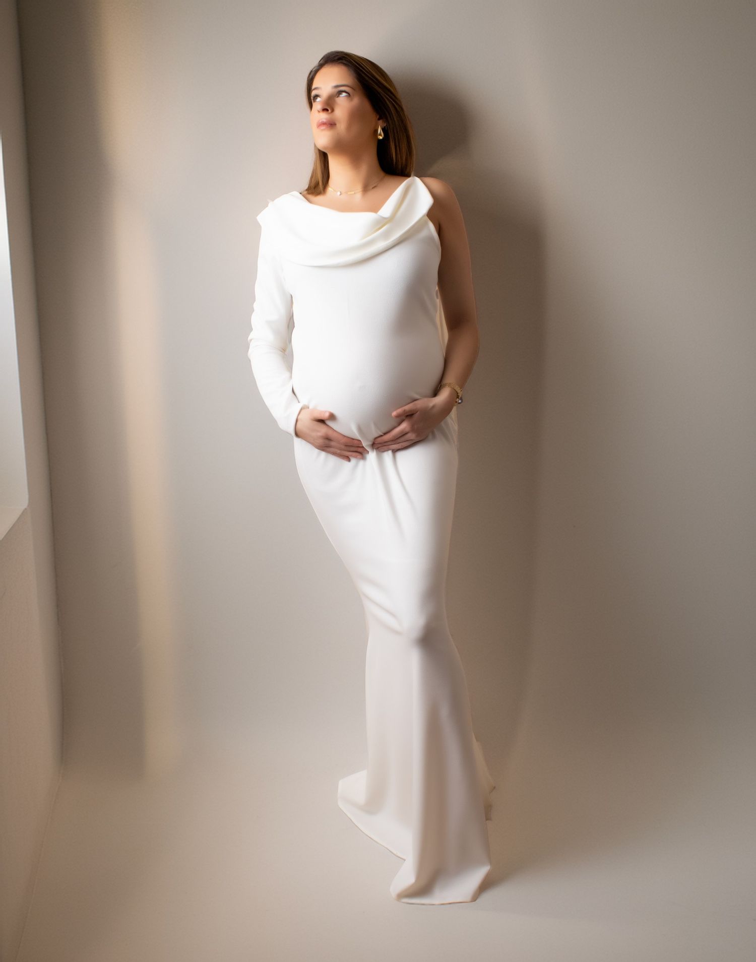 Pregnant woman in flowing white maternity gown gently holding baby bump against neutral wall.