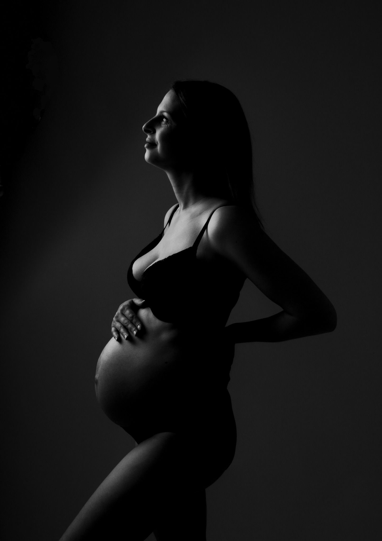 Dramatic black and white silhouette maternity portrait of pregnant woman in lingerie against dark background.