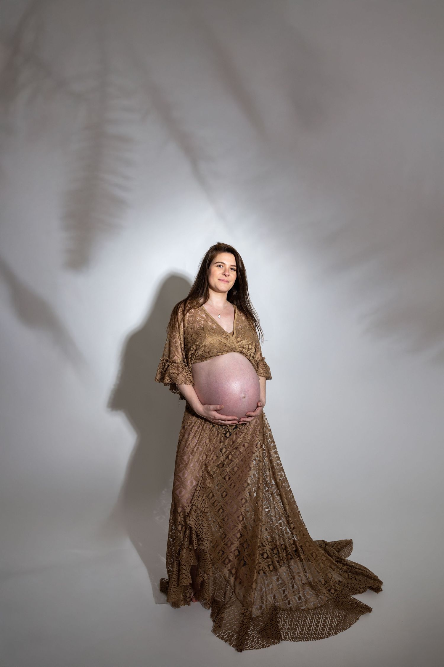 Pregnant woman in elegant gold lace dress cradling her baby bump in a studio with soft fern shadow backdrop.