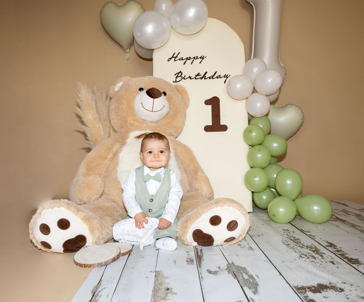 Smiling baby boy in white suit sits with giant teddy bear at first birthday balloon backdrop.