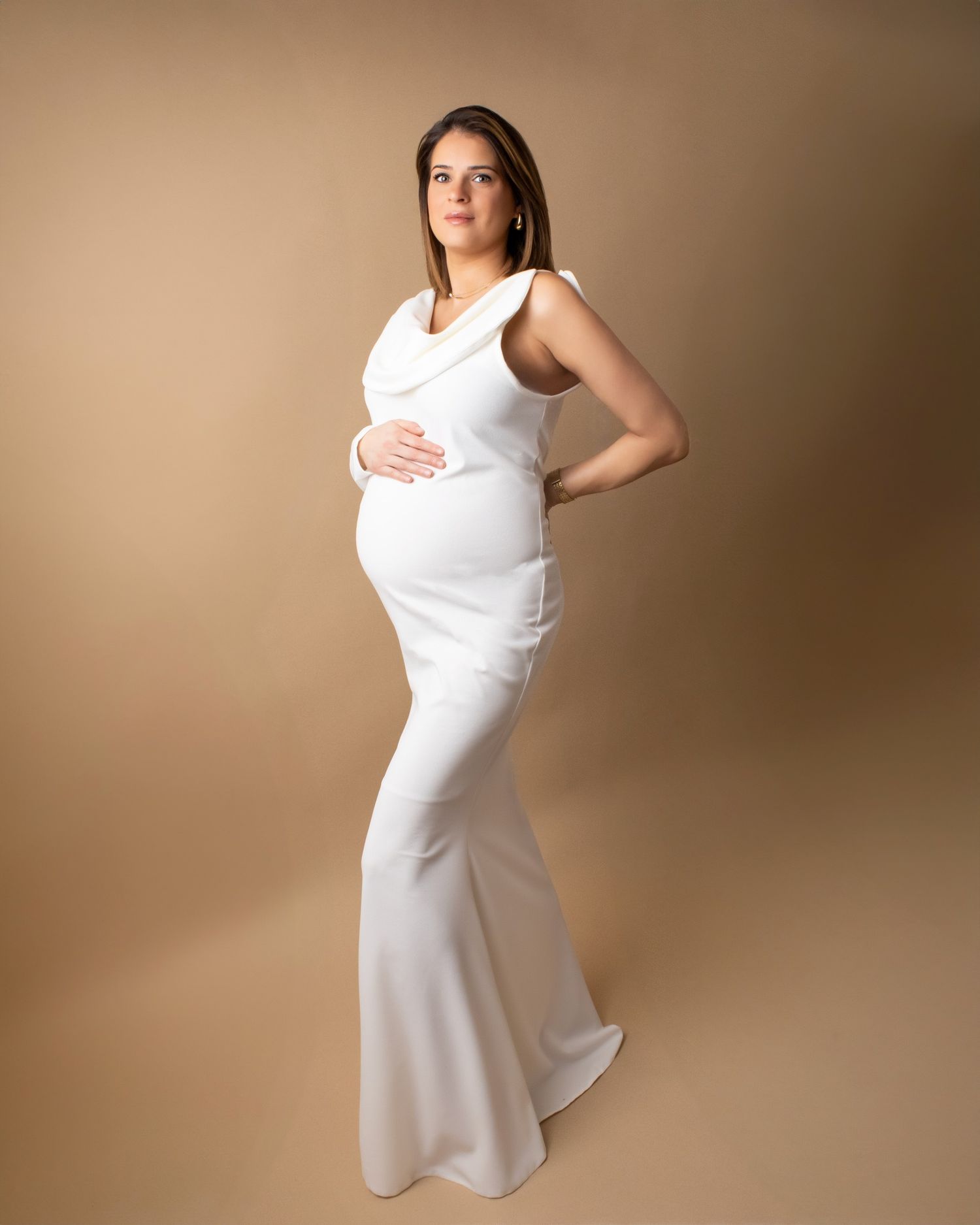 Pregnant woman in sleeveless white fitted maternity gown posing sideways on beige studio background.