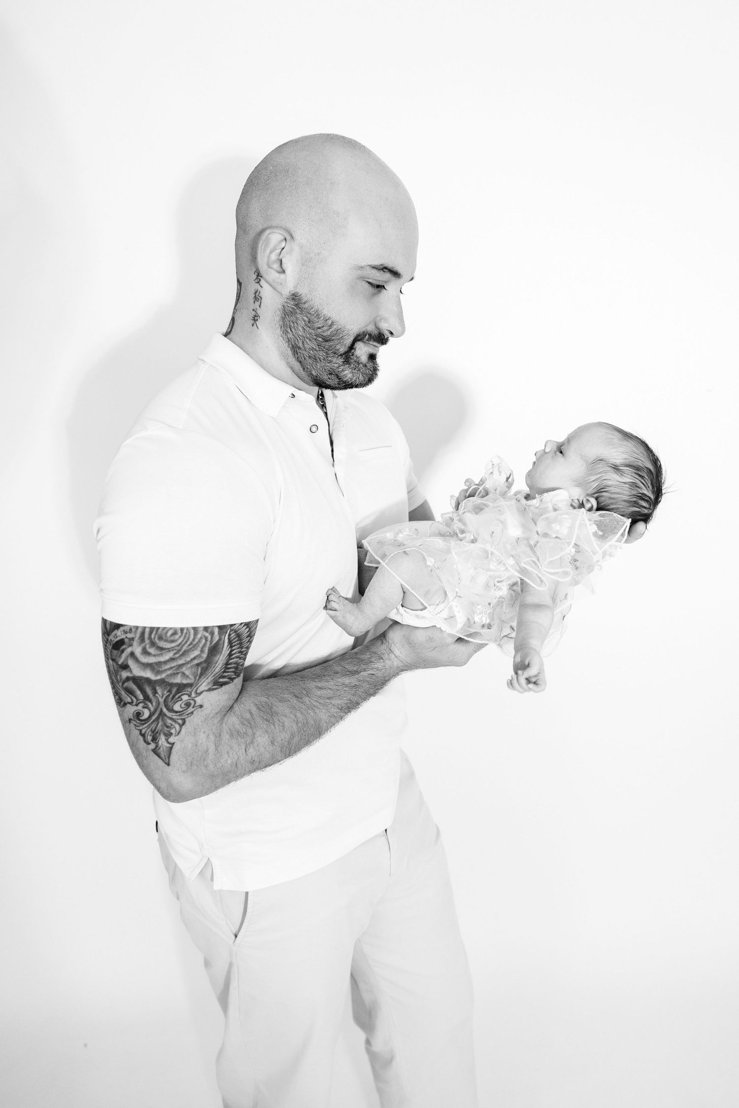 A bald tattooed father lovingly cradles his newborn baby in a black and white studio portrait.