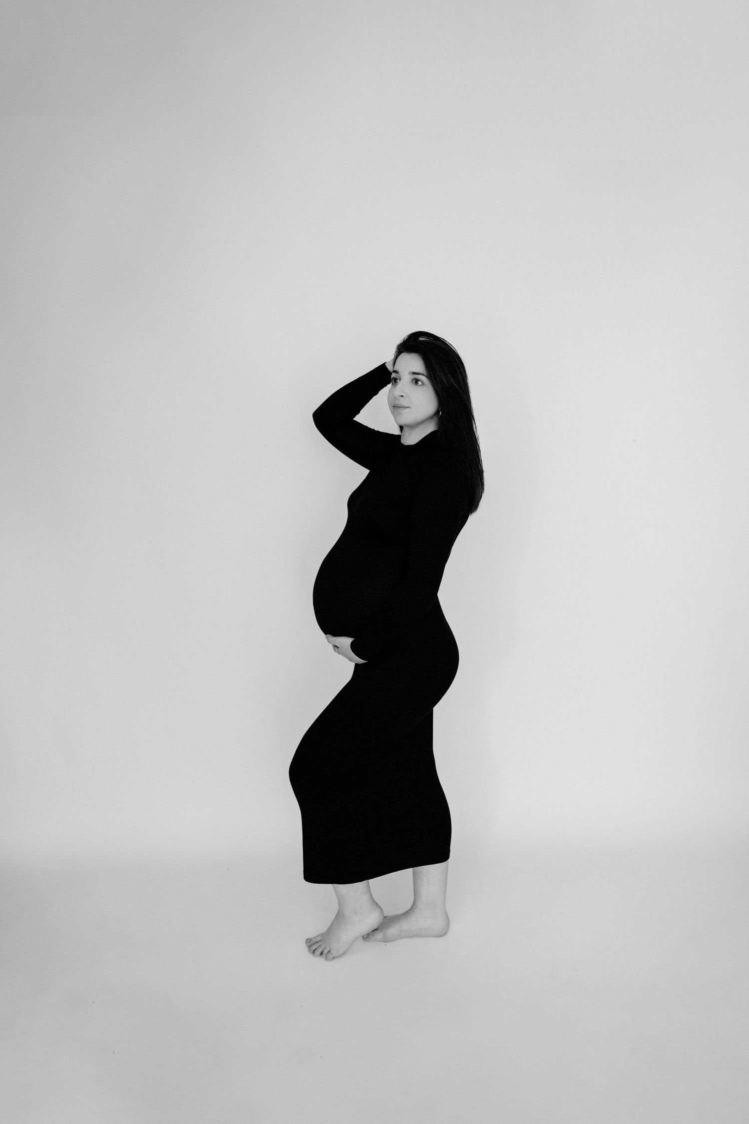 Black and white maternity photo of a pregnant woman in a black dress, posing barefoot against a white background.