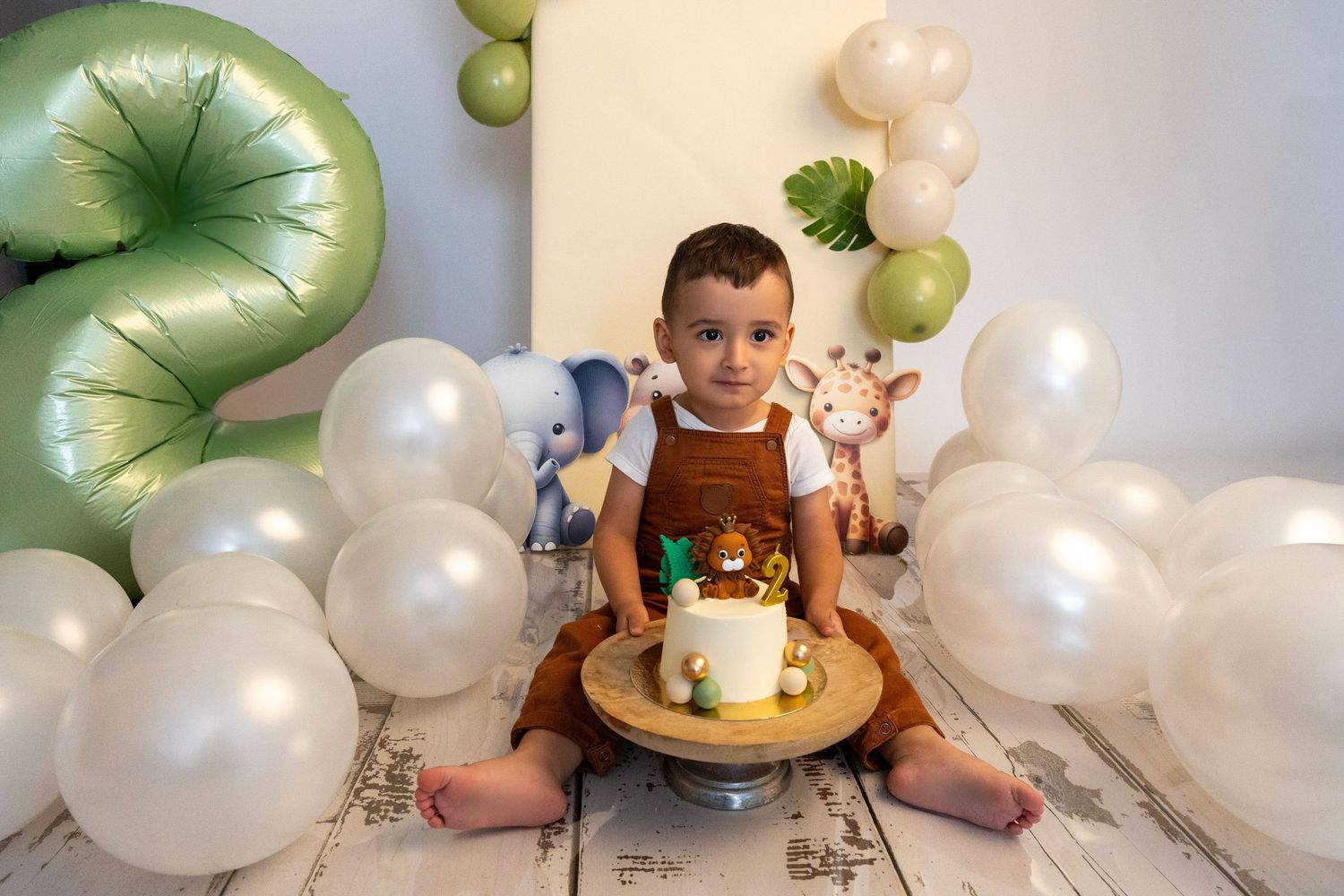 Baby boy in brown overalls sitting with a safari-themed cake surrounded by white and green balloons.