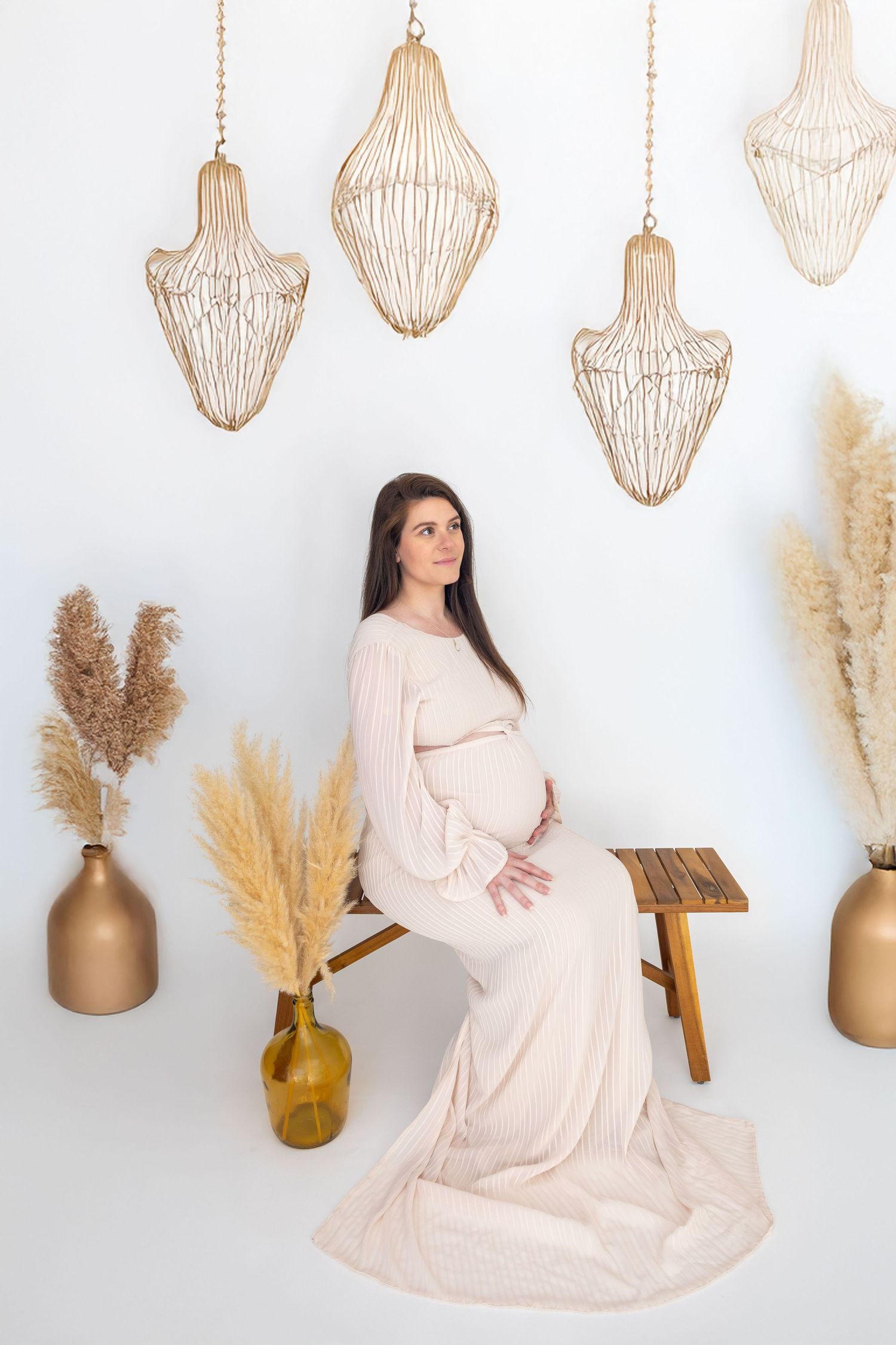 Pregnant woman in blush gown sits on wooden bench surrounded by pampas grass and gold wire chandeliers in boho studio.