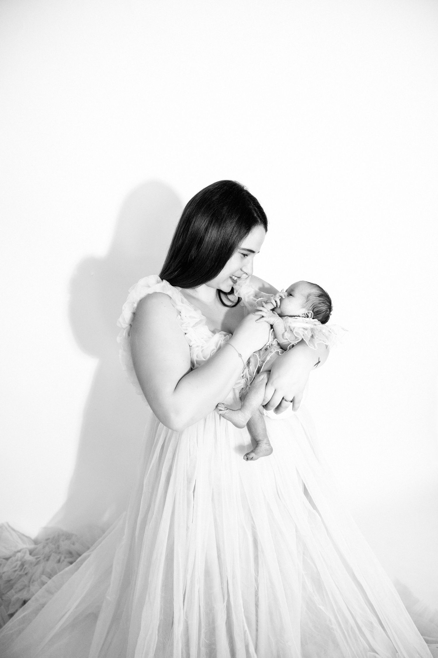 A mother in a flowing white gown tenderly holds her newborn baby in a black and white portrait.