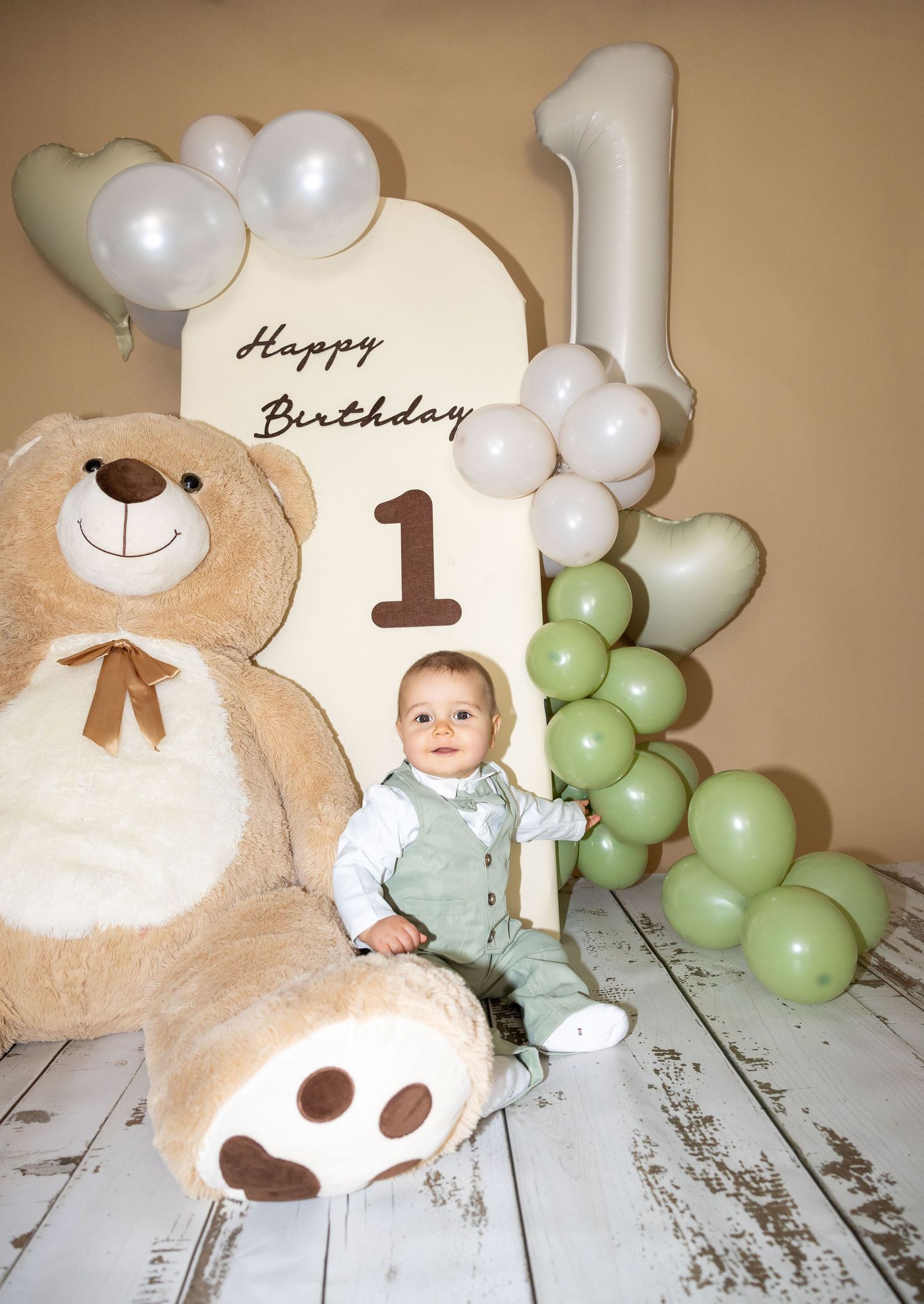 Baby boy in green outfit sits beside giant teddy bear at first birthday balloon celebration.