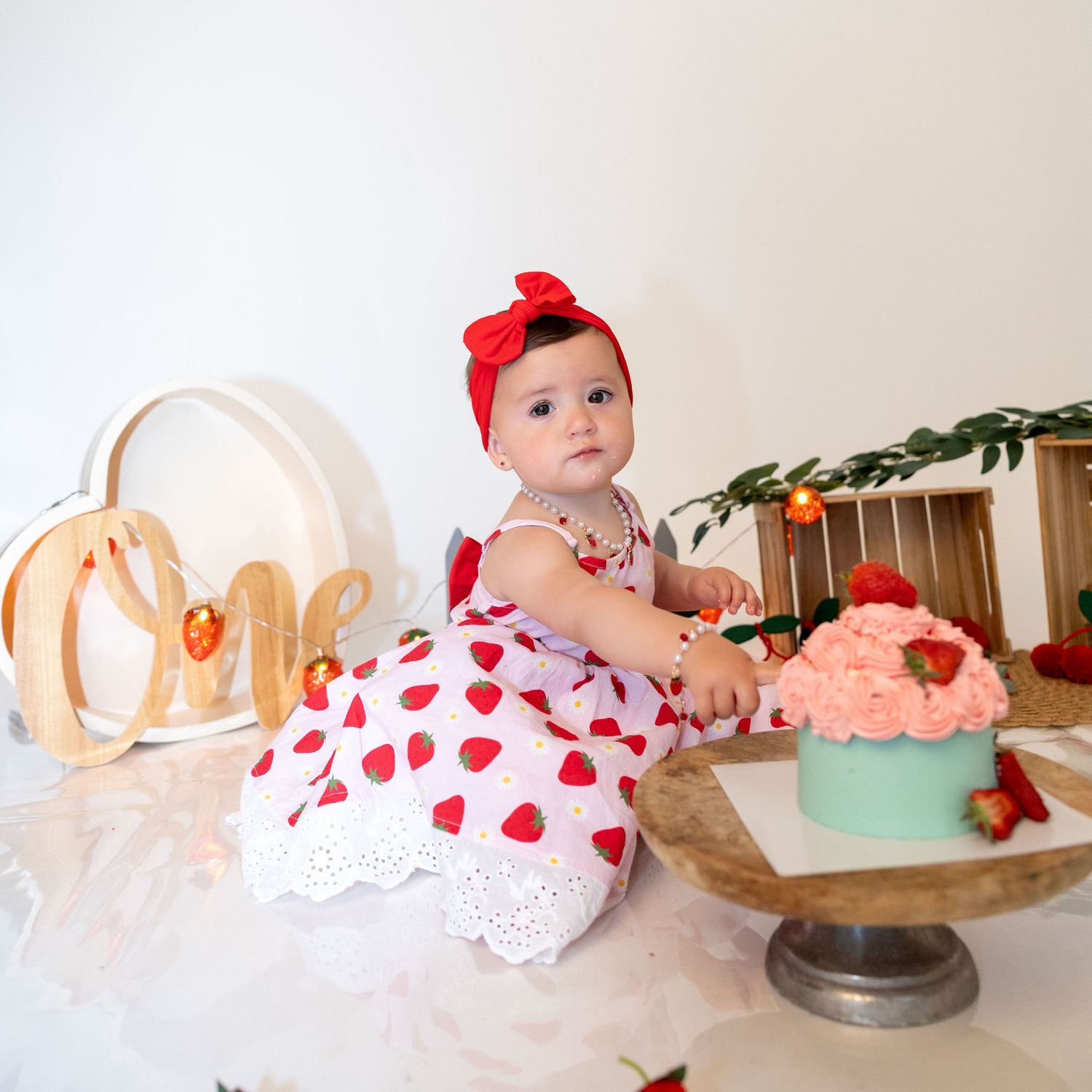 A baby girl in a heart-print dress reaches toward a pink cake during her first birthday session.
