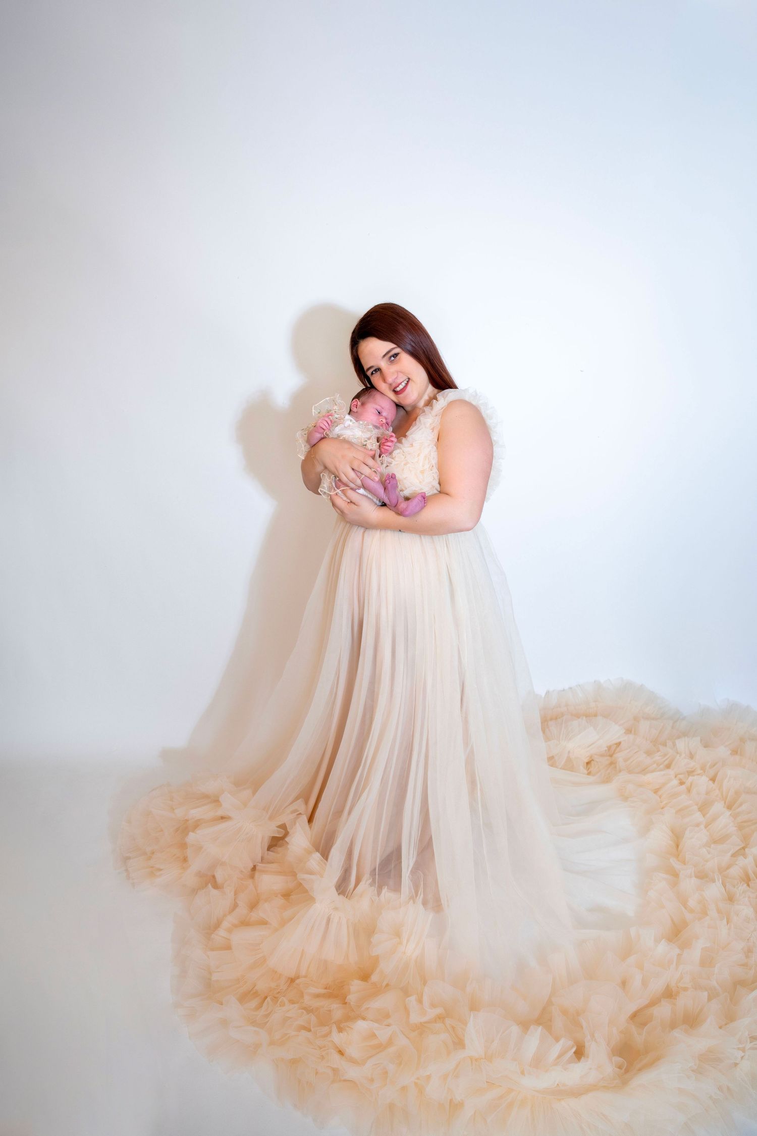 A mother in an elegant champagne tulle gown smiles while holding her newborn baby in a studio.