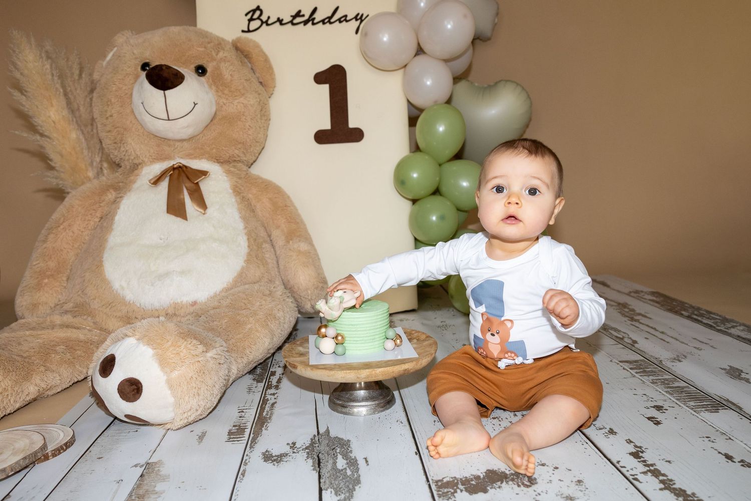 A baby celebrates their first birthday with a cake and large teddy bear, wearing a bear-print outfit.
