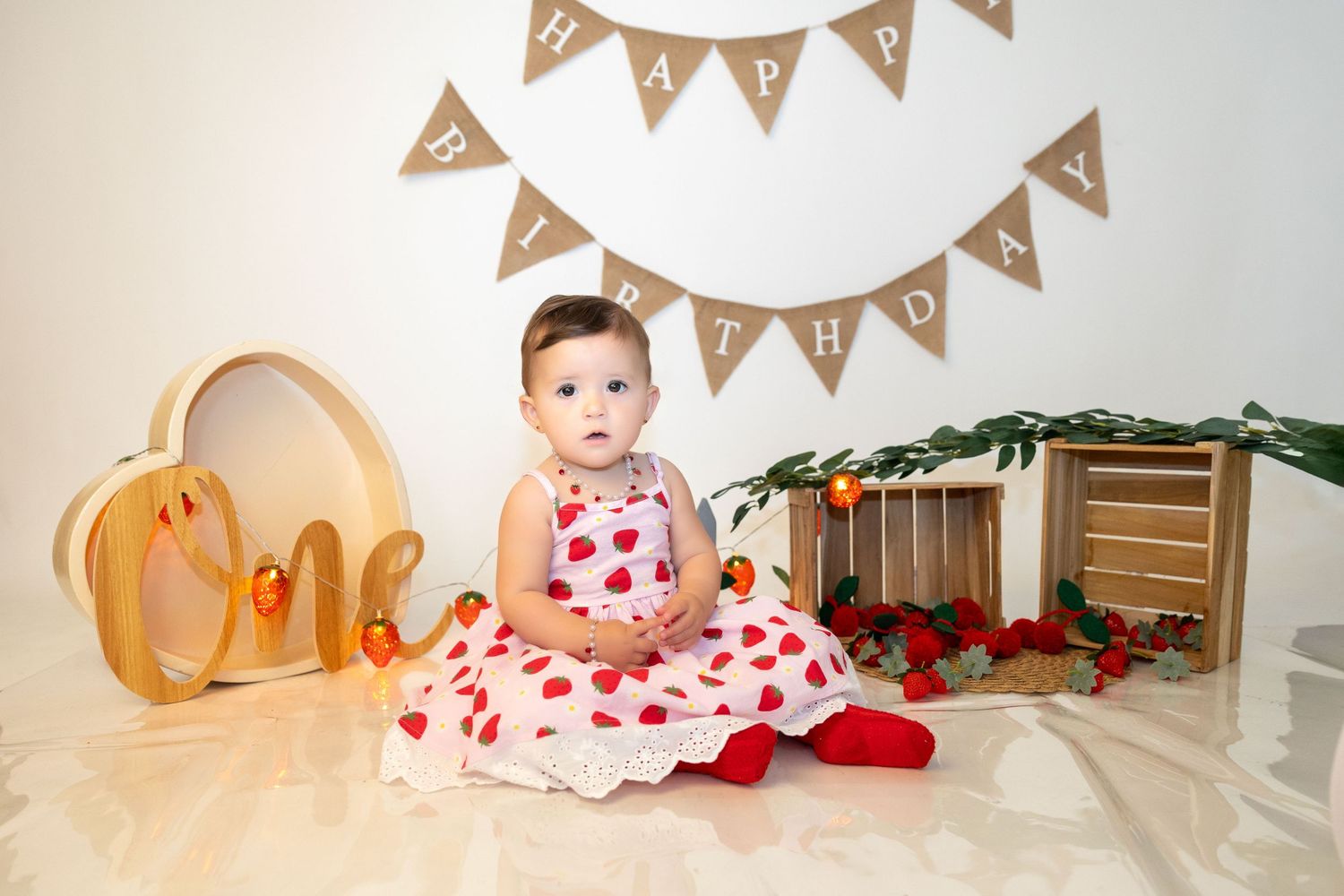 A baby girl in a heart-print dress sits near a Happy Birthday banner during her first birthday.