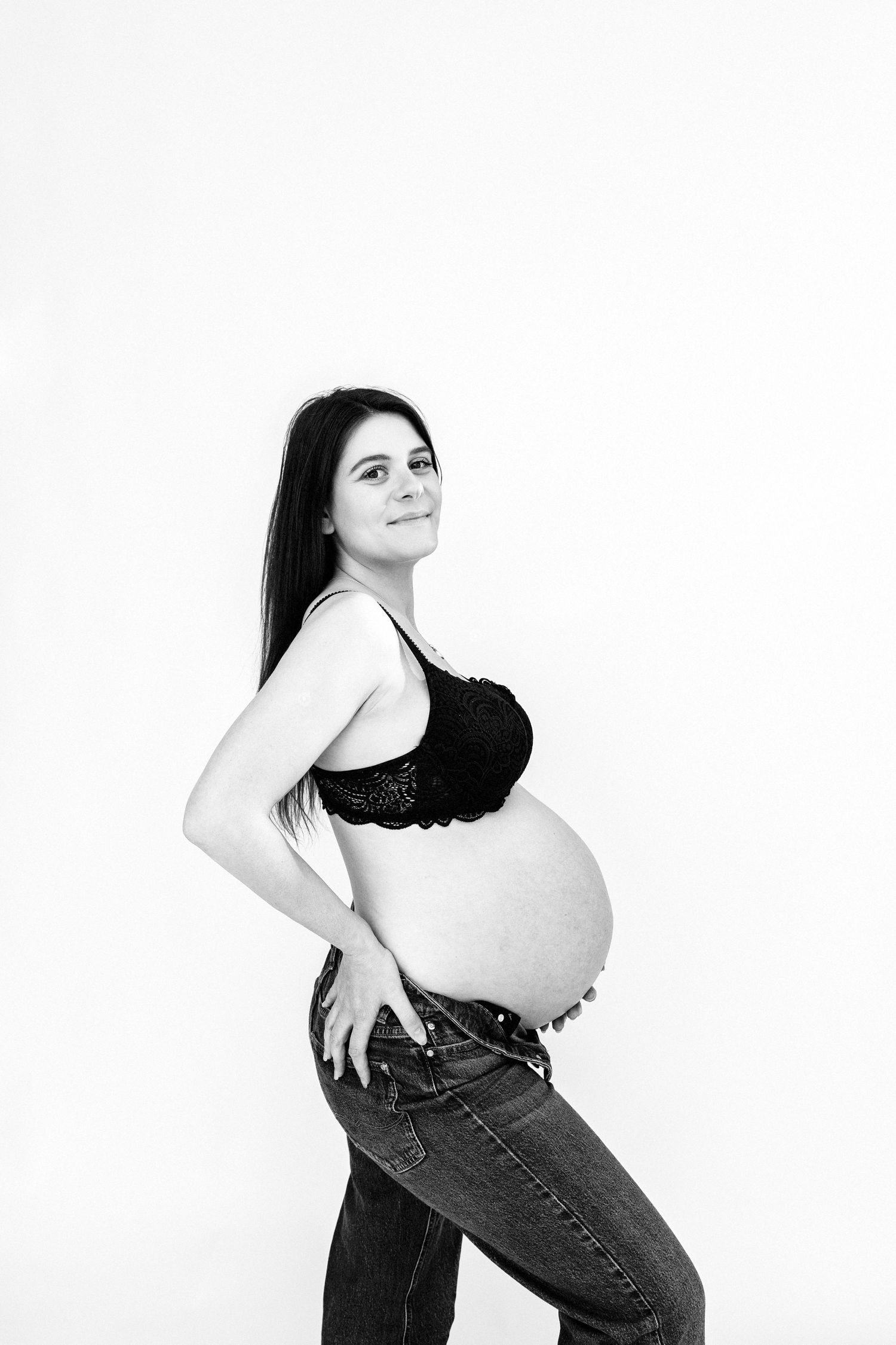 Black and white portrait of a smiling pregnant woman in a black bralette and jeans showcasing her baby bump.