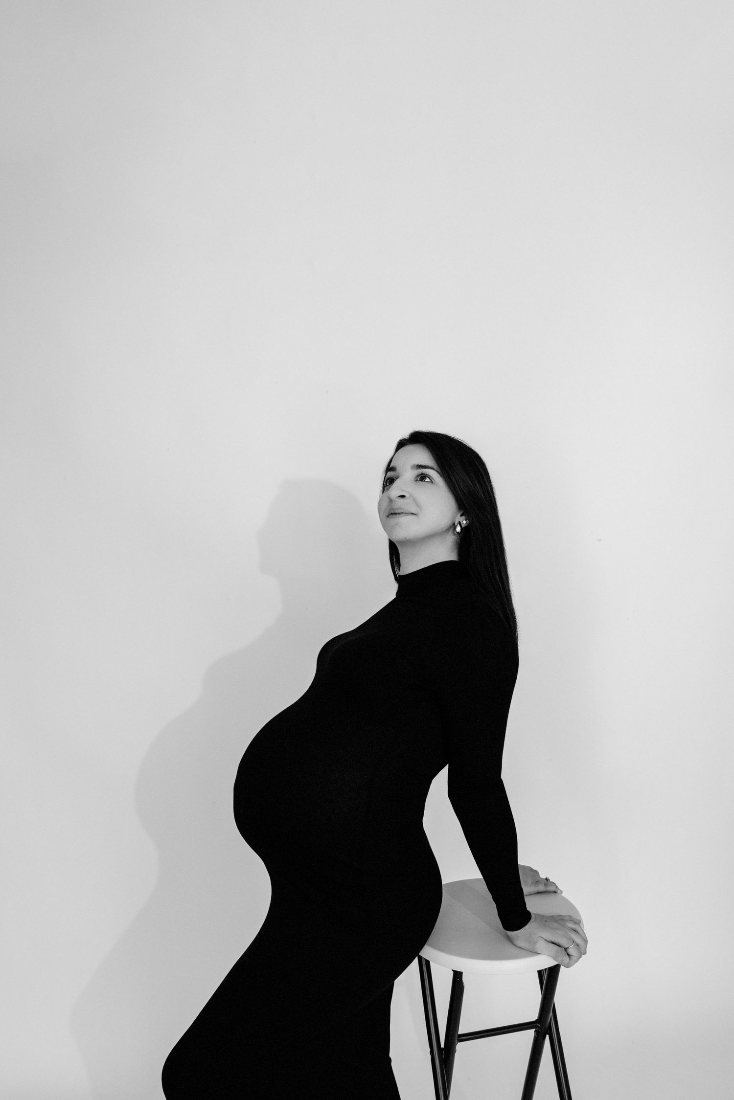 Black and white portrait of a pregnant woman in a black dress sitting on a stool, looking upward gracefully.
