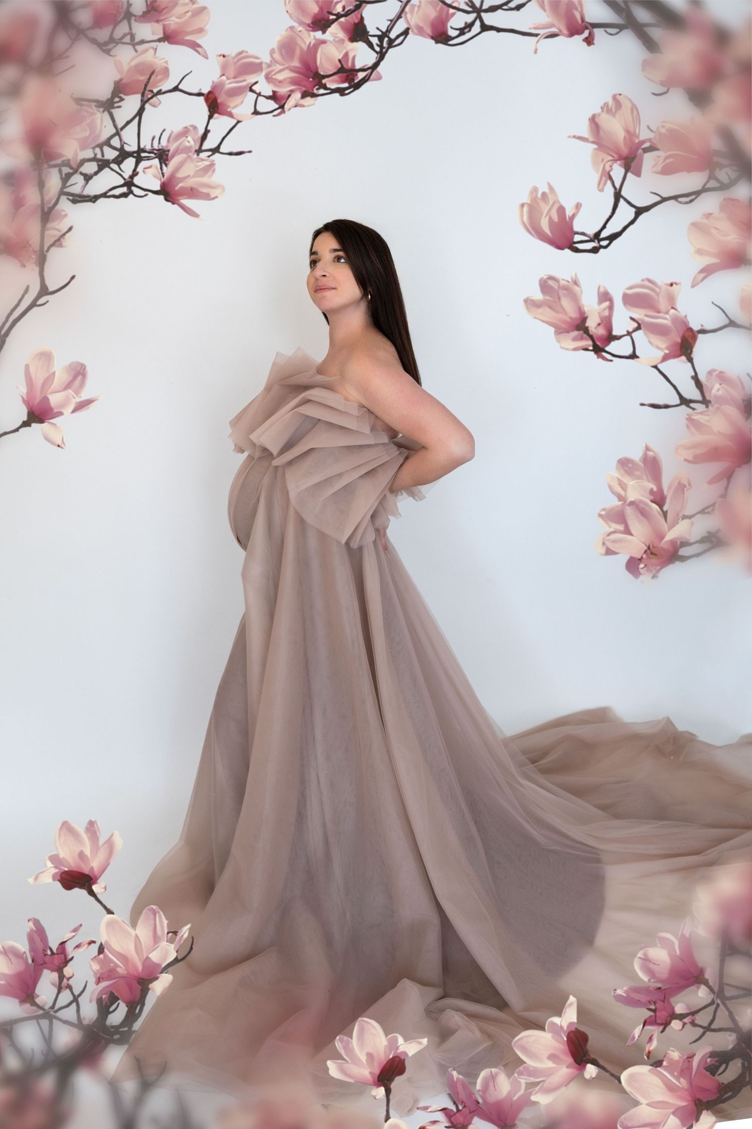 Pregnant woman in elegant taupe tulle gown surrounded by pink magnolia blossoms in a dreamy maternity portrait.