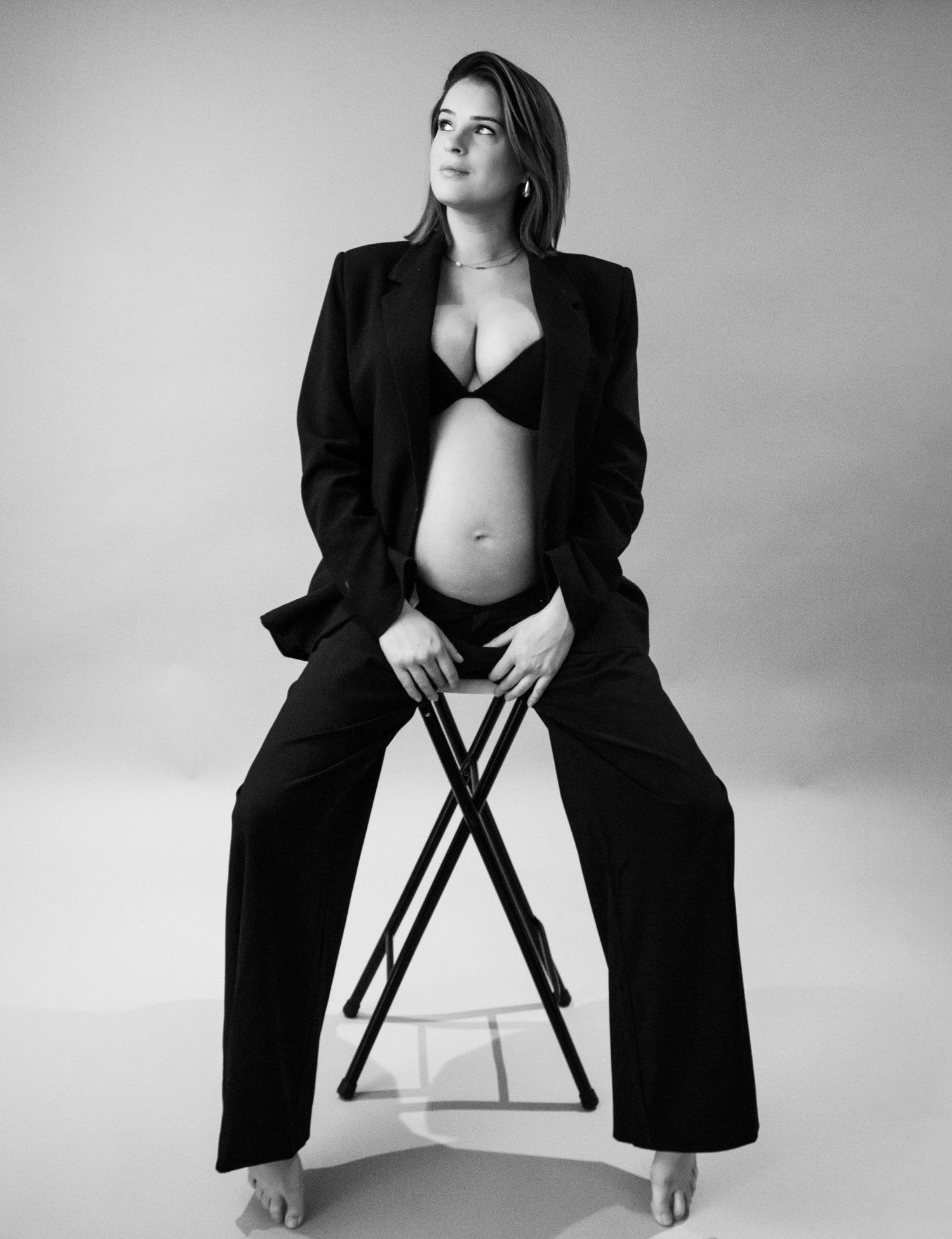 Pregnant woman in black blazer and bra sitting on stool in black and white studio portrait.