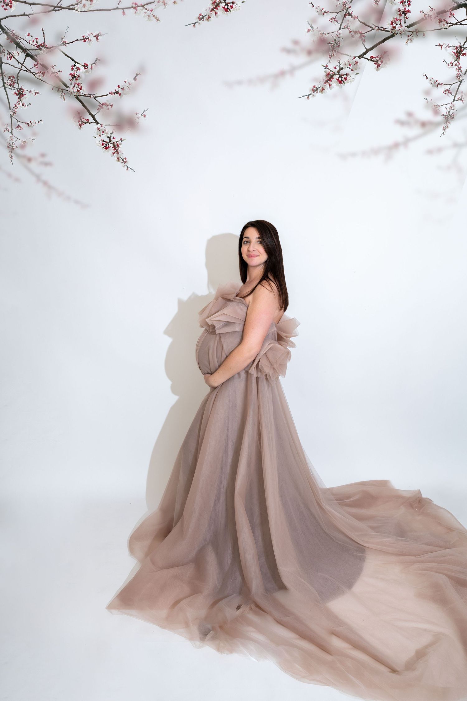 Pregnant woman in flowing taupe tulle gown poses gracefully under cherry blossom branches in a bright studio setting.