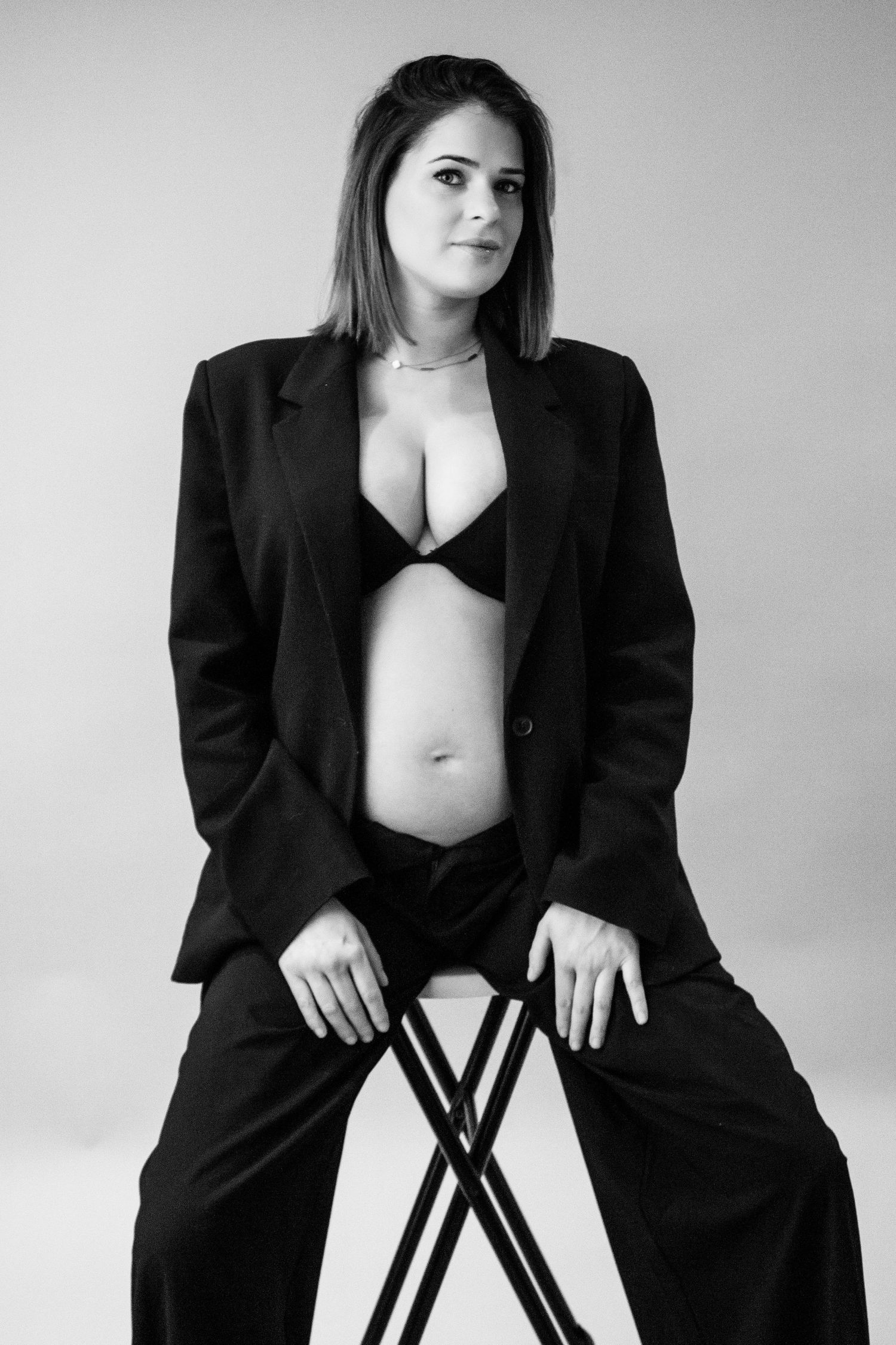 Pregnant woman in black blazer and bra sitting on stool looking at camera in monochrome portrait.