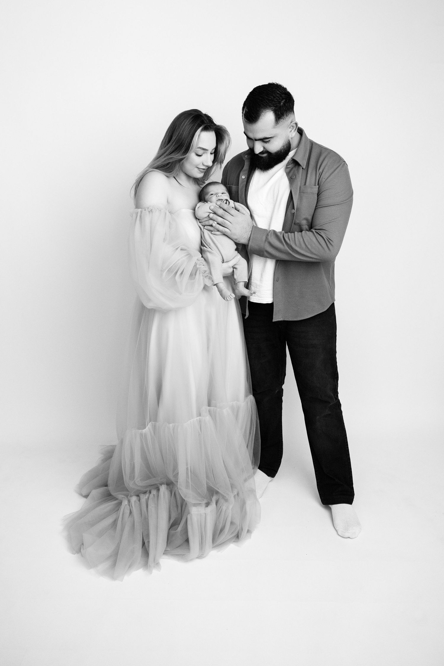 A couple in a flowing tulle gown tenderly gazes at their newborn baby in a black and white portrait.