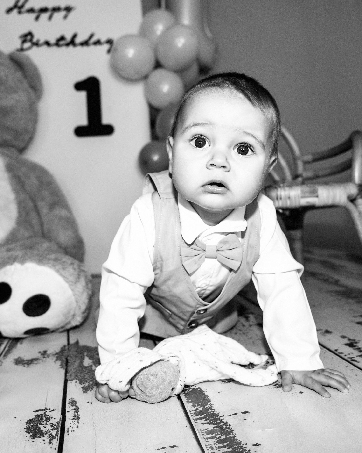 Black and white portrait of a baby boy in bow tie vest crawling at first birthday party.
