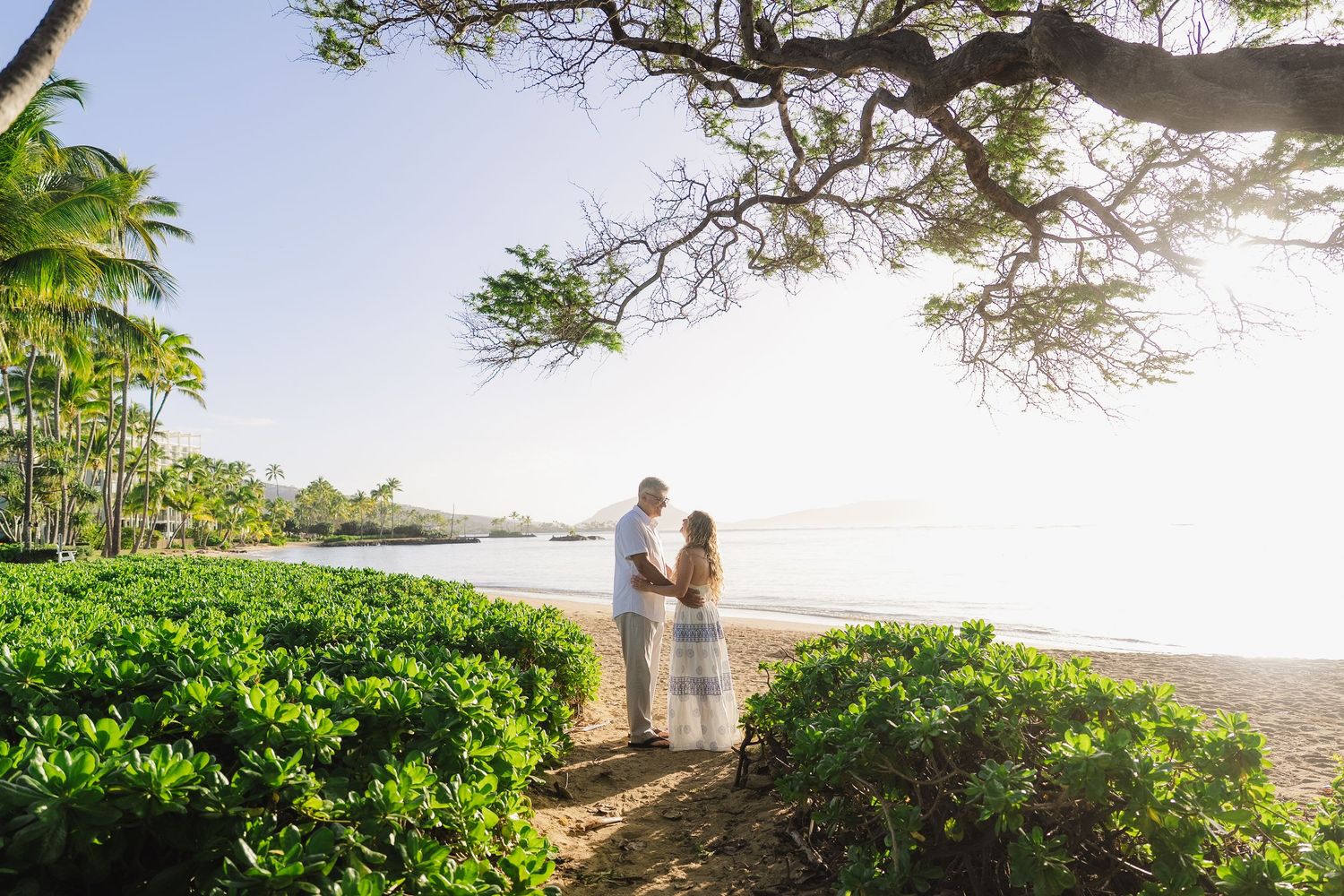 A romantic beach wedding ceremony setup with white chairs and floral arch overlooking the ocean at sunset.