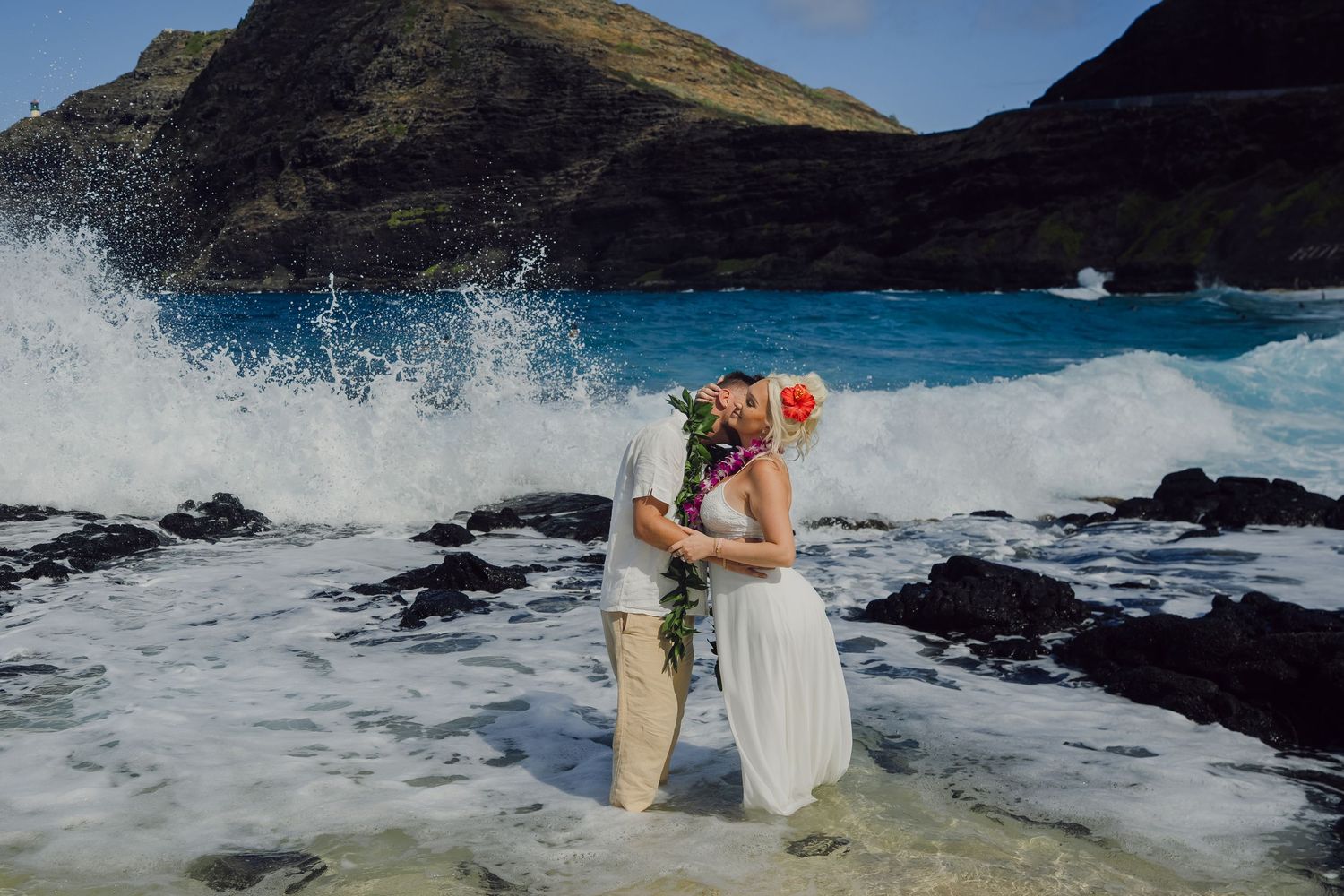 Couple kissing on rocky Hawaii shore with surfboard, floral lei, white dress, and crashing ocean waves against green mountains.