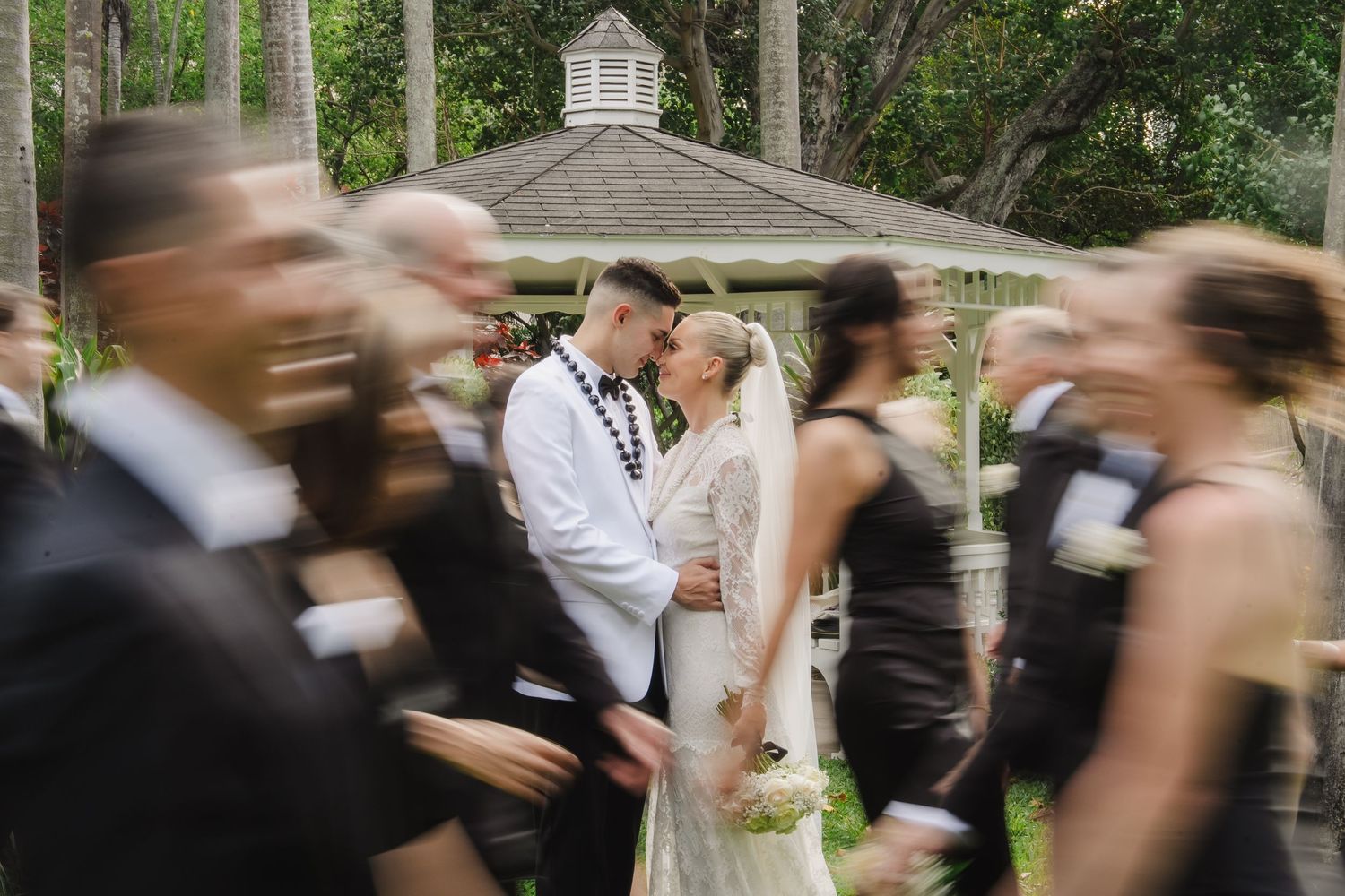 Wedding party members walk in a processional line outdoors between rows of chairs during a garden ceremony.