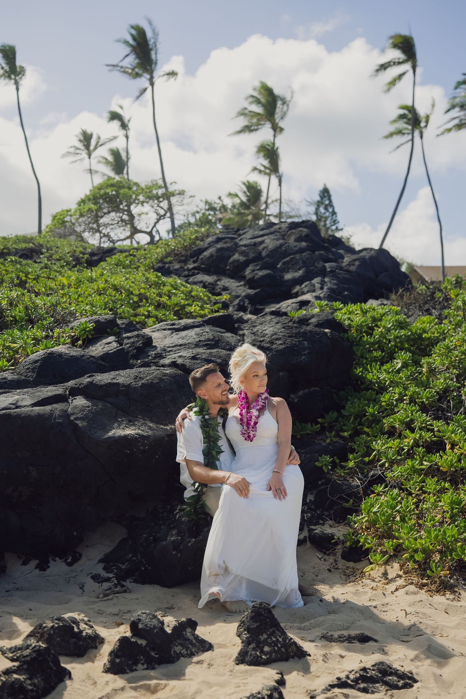 Bride and groom in leis embrace on a sandy Hawaiian beach with black lava rocks and swaying palm trees.