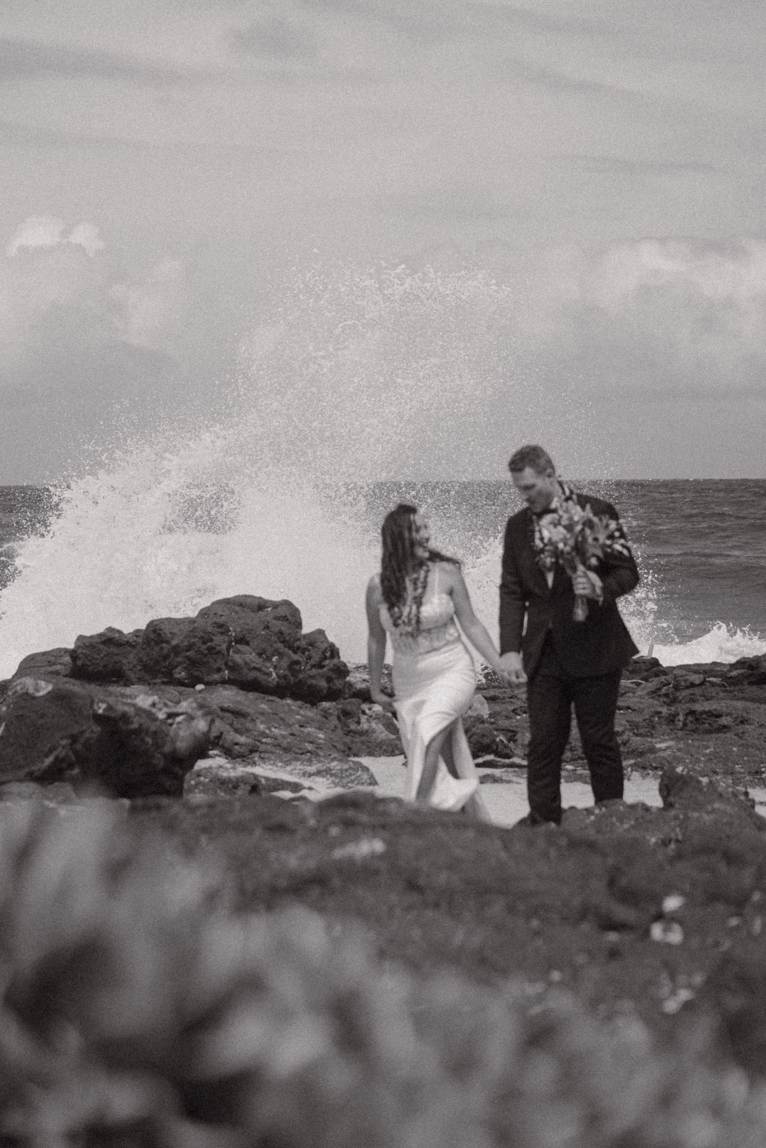 A couple in wedding attire stands on rocky coastal shore as dramatic waves crash behind them in black and white.