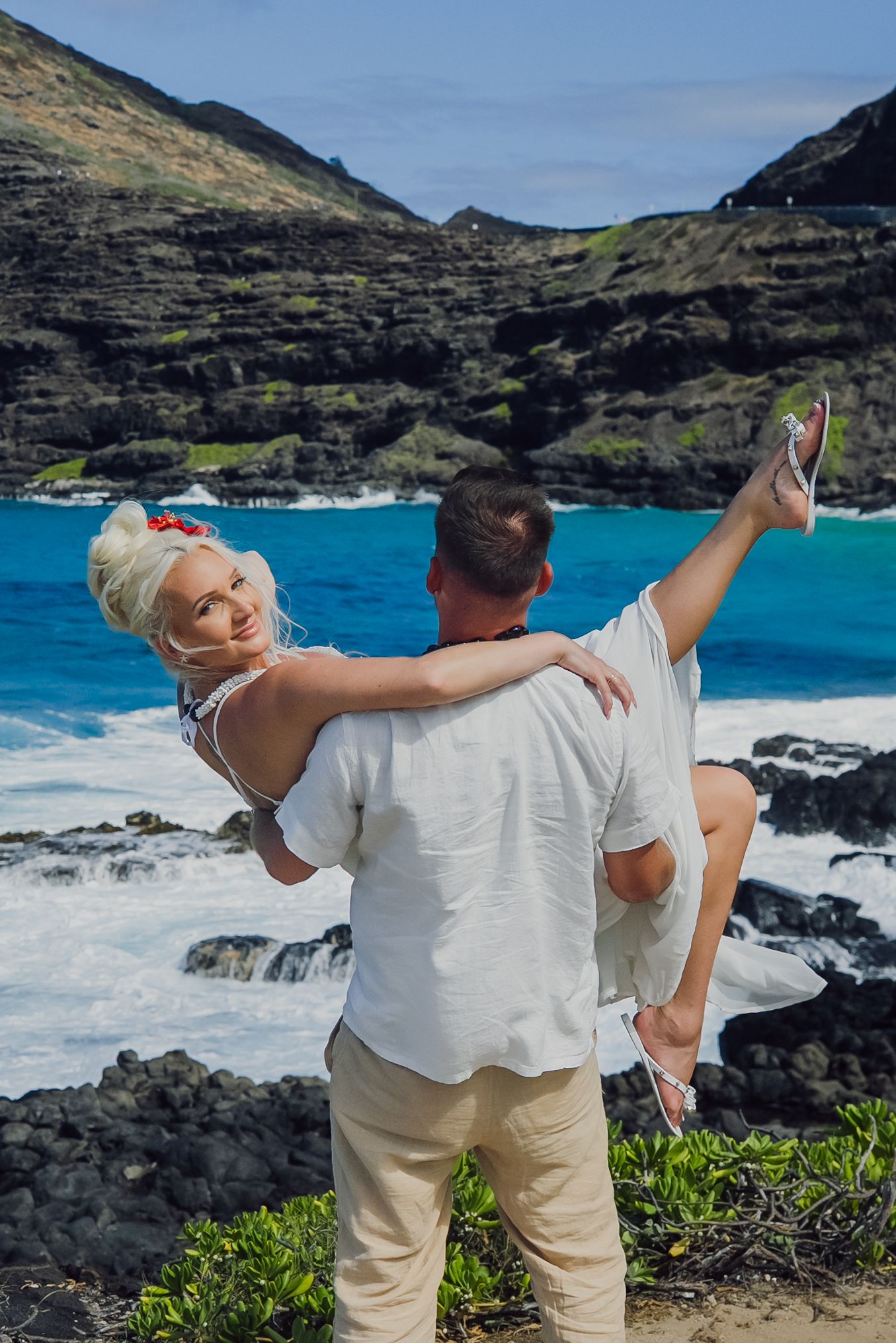 Couple embracing on rocky Hawaiian coastline, man lifting woman in white dress with turquoise ocean waves and green mountains behind.