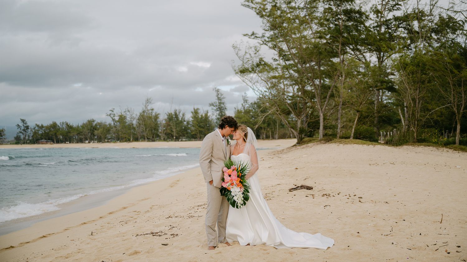 Bride and groom kissing on a tropical beach with lush trees and ocean waves, bride holding a pink bouquet.