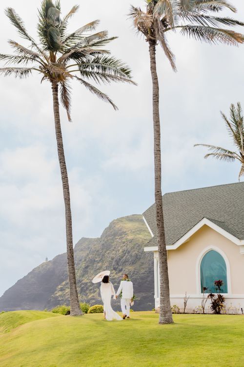 Couple in white attire walking on lush green lawn with tall palm trees and a chapel near Hawaii mountains.