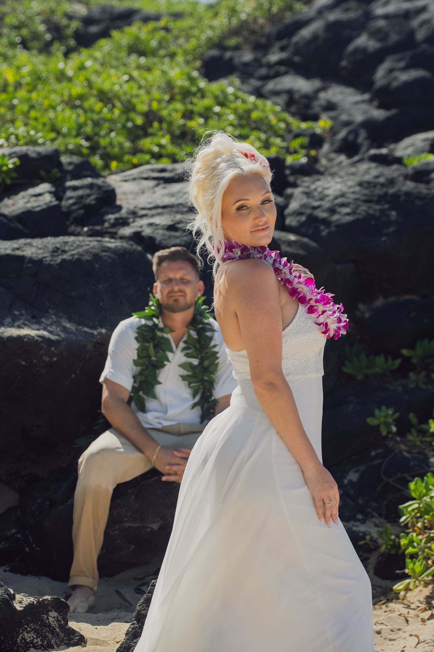 Bride in white dress and pink lei poses with groom in white shirt and green lei on rocky Hawaiian beach.