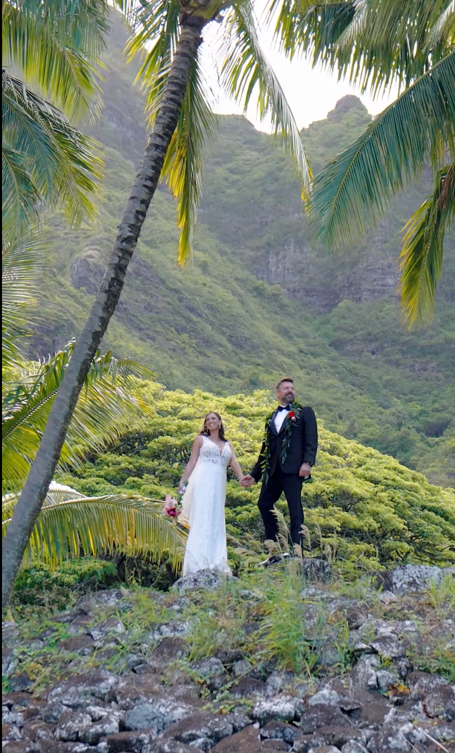 Bride and groom hold hands on rocky hillside with lush green mountains and palm trees in tropical Hawaii wedding.
