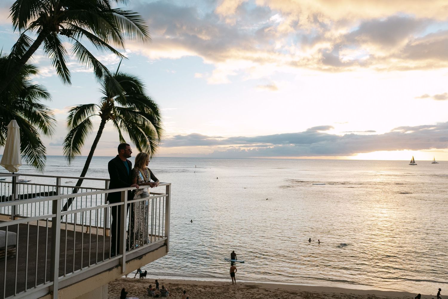 Silhouetted palm trees frame a golden sunset view from a beachfront balcony in Hawaii with ocean and mountains in the distance.
