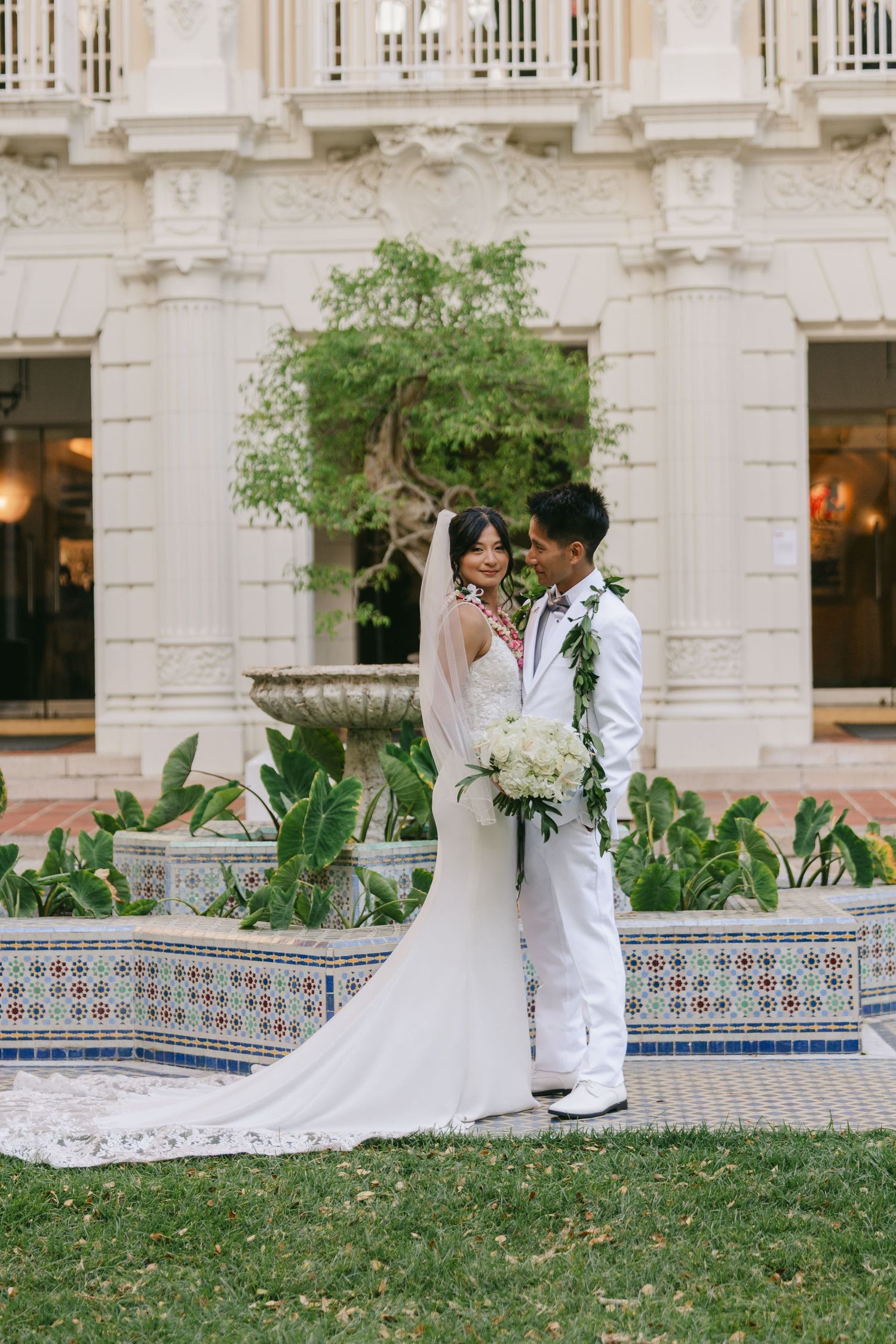 Newlyweds share an intimate moment in front of an ornate building with tiled fountain and lush greenery.