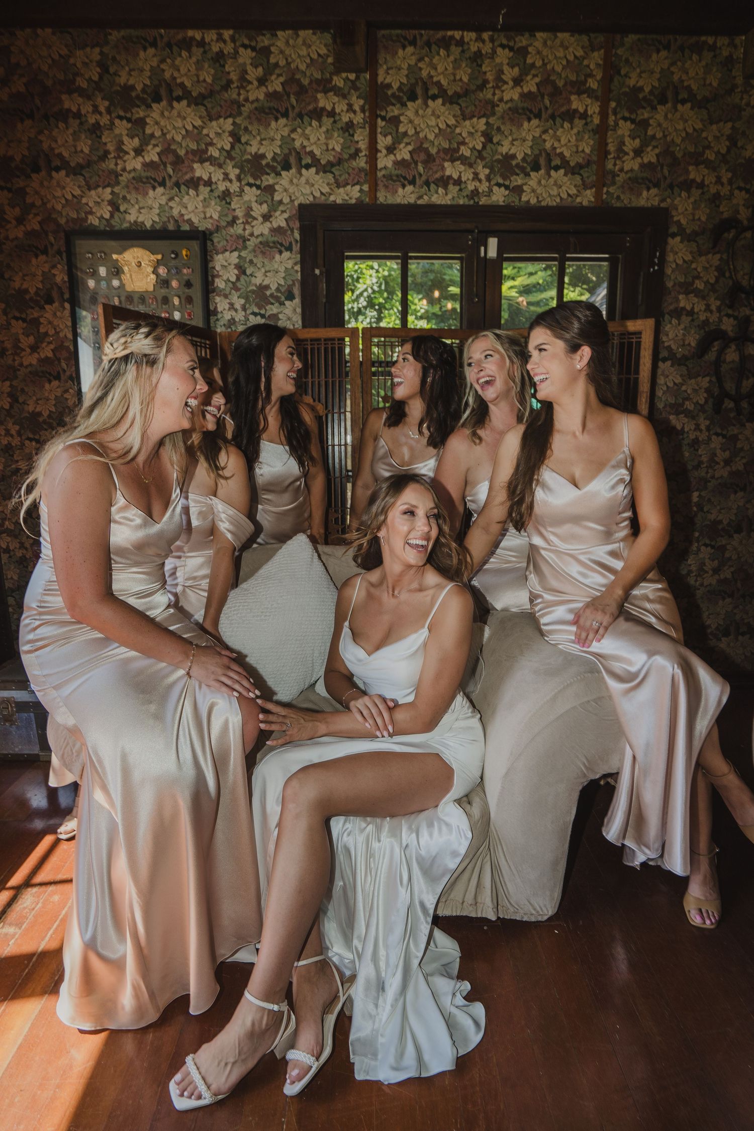 A group in ivory satin dresses gather by a window in a rustic room with natural lighting streaming in.