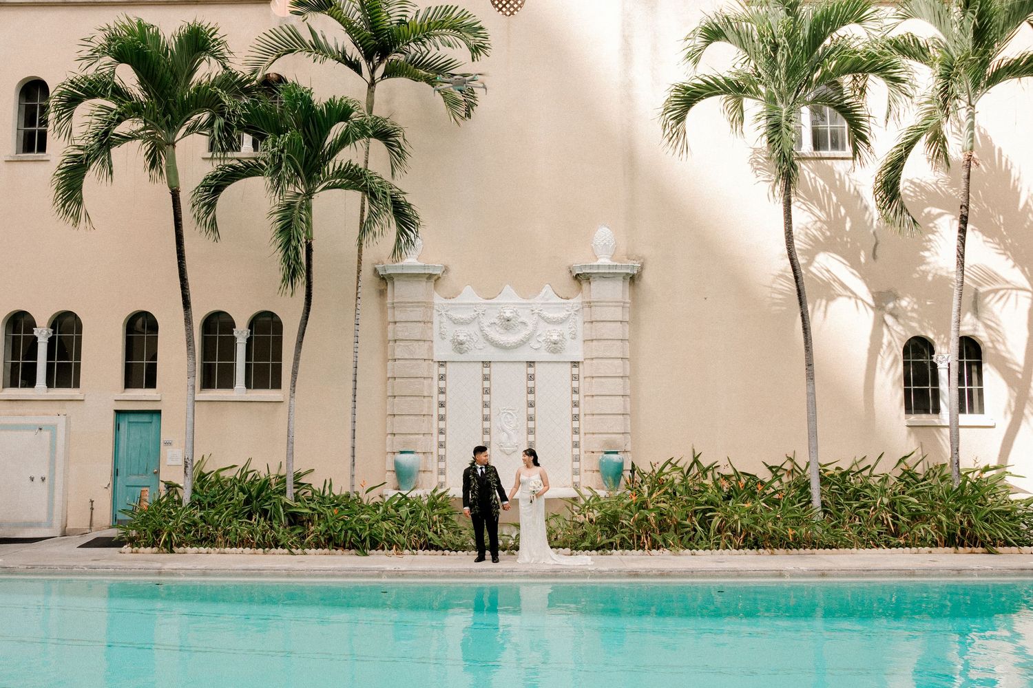 Bride and groom stand by a pool with palm trees and elegant building backdrop at a luxury wedding venue.