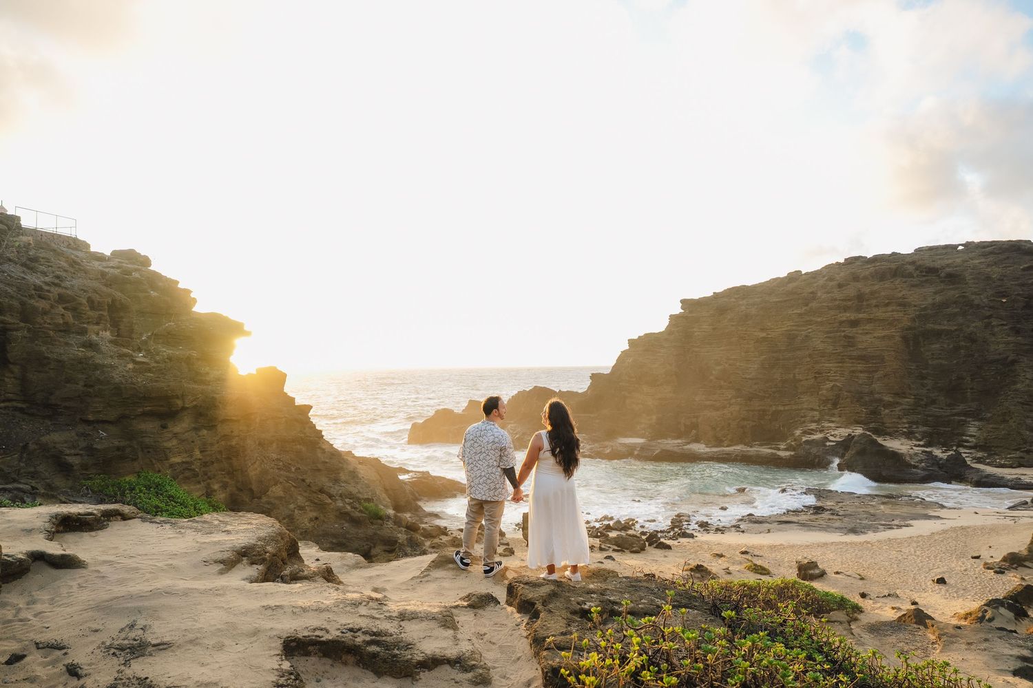 A couple in white stands on rocky cliffs overlooking the ocean at sunset, surrounded by rugged coastal scenery.