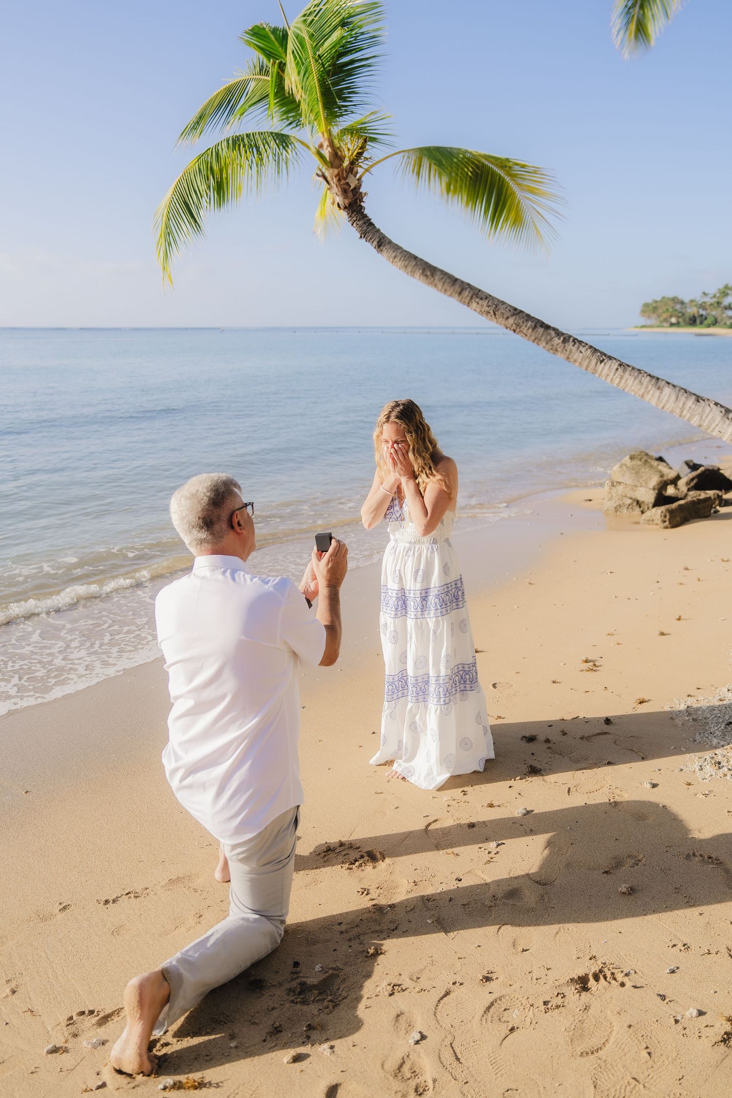 Couple in white clothing posing for romantic photos under palm trees on a tropical beach at sunset.