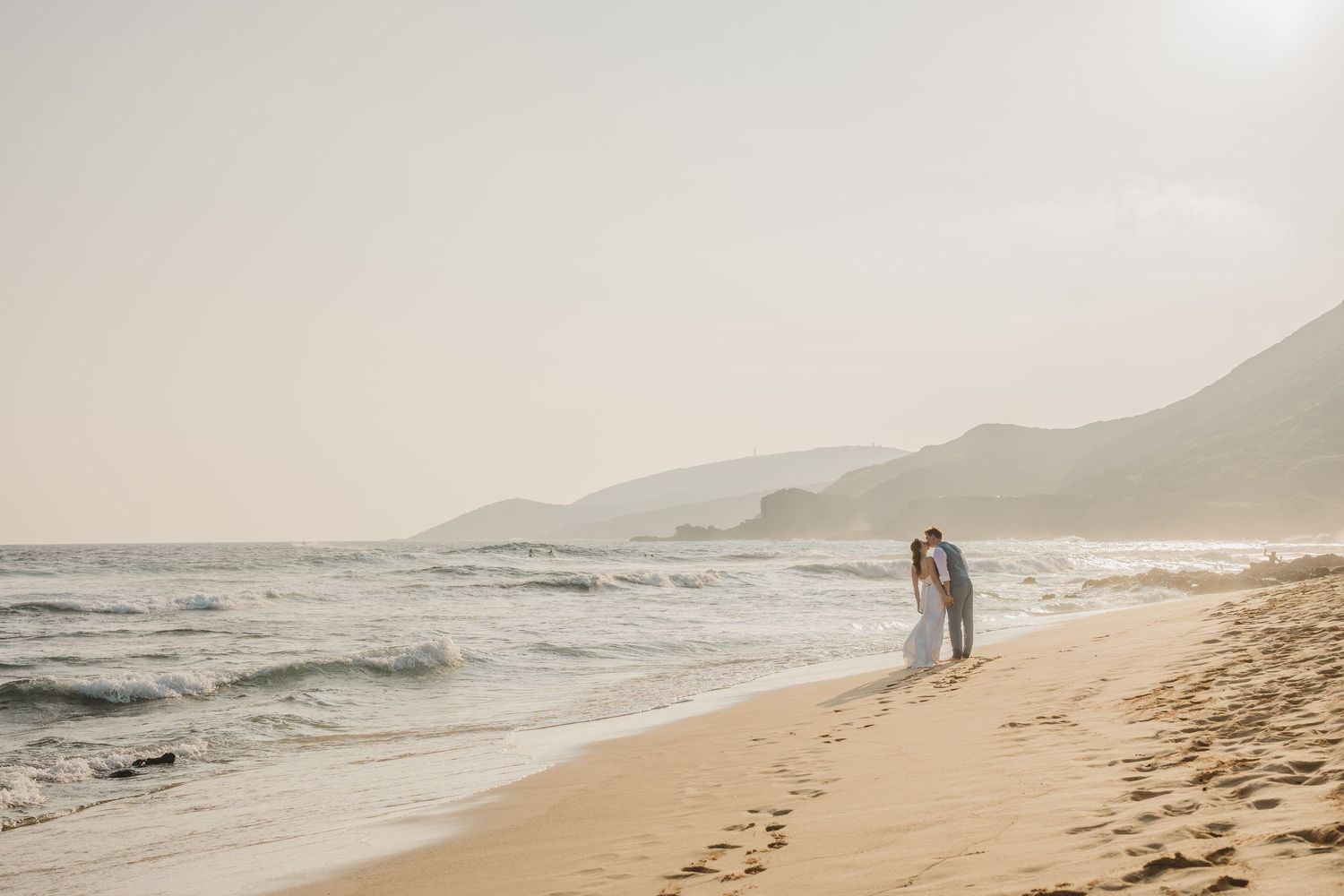 Couple walking along misty beach shoreline with mountains in the background on an overcast day.